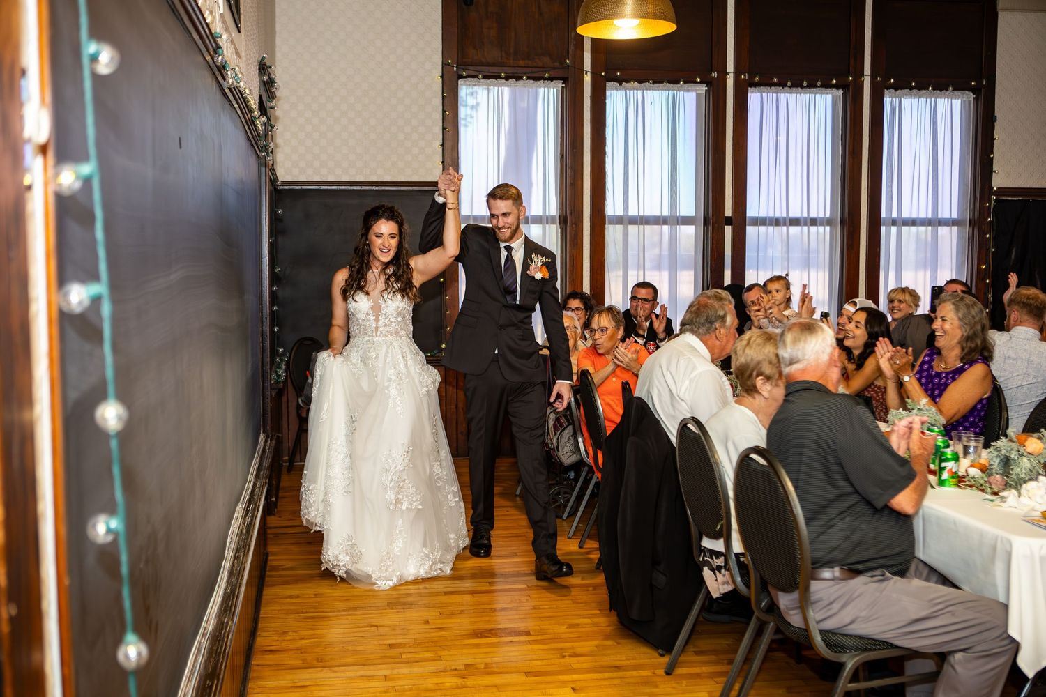 Bride and groom make grand entrance at wedding reception, guests applauding in elegant wood-floored venue.