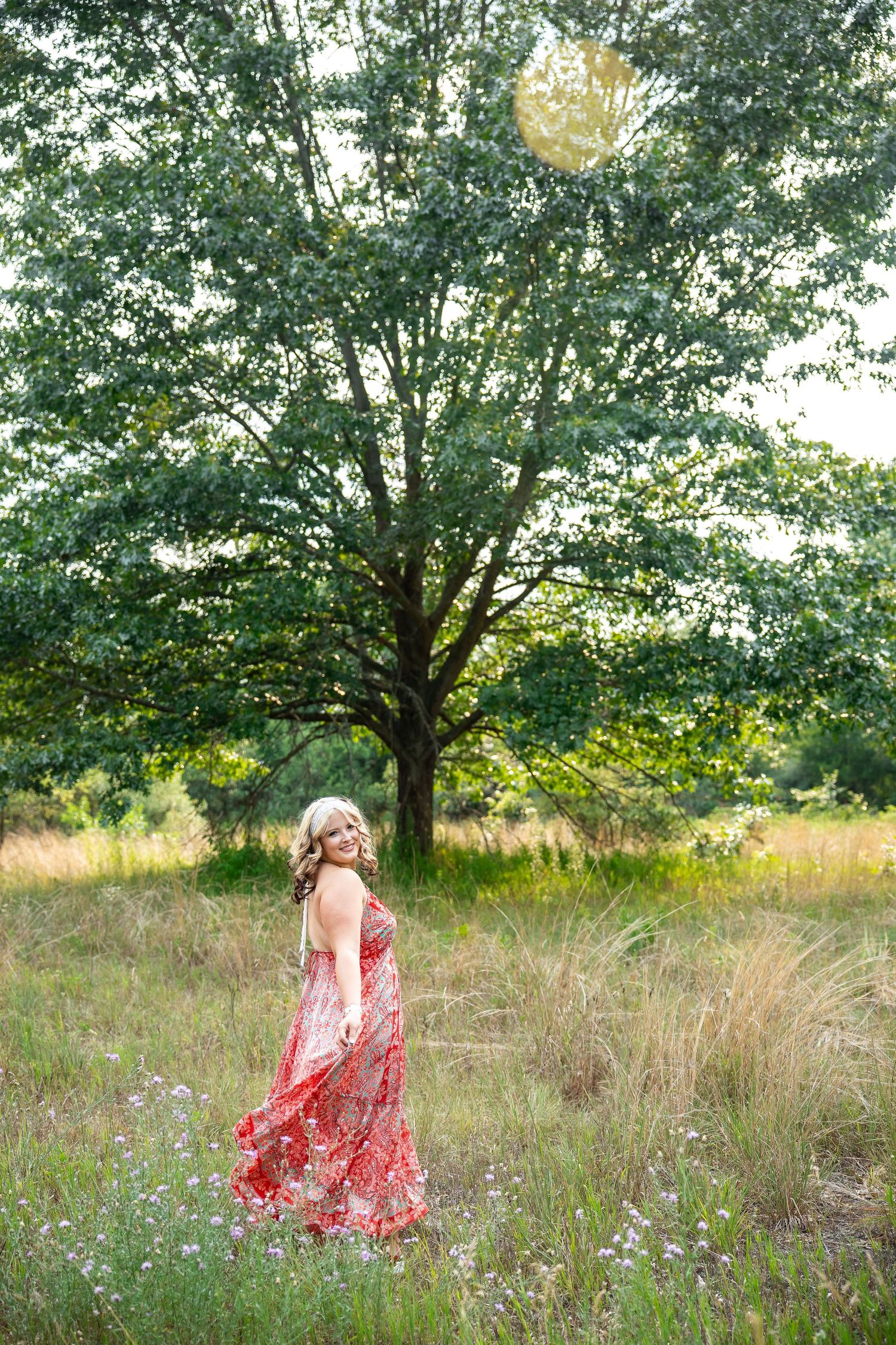 A smiling woman in a flowing red floral maxi dress stands in a lush green meadow with a large tree behind her.