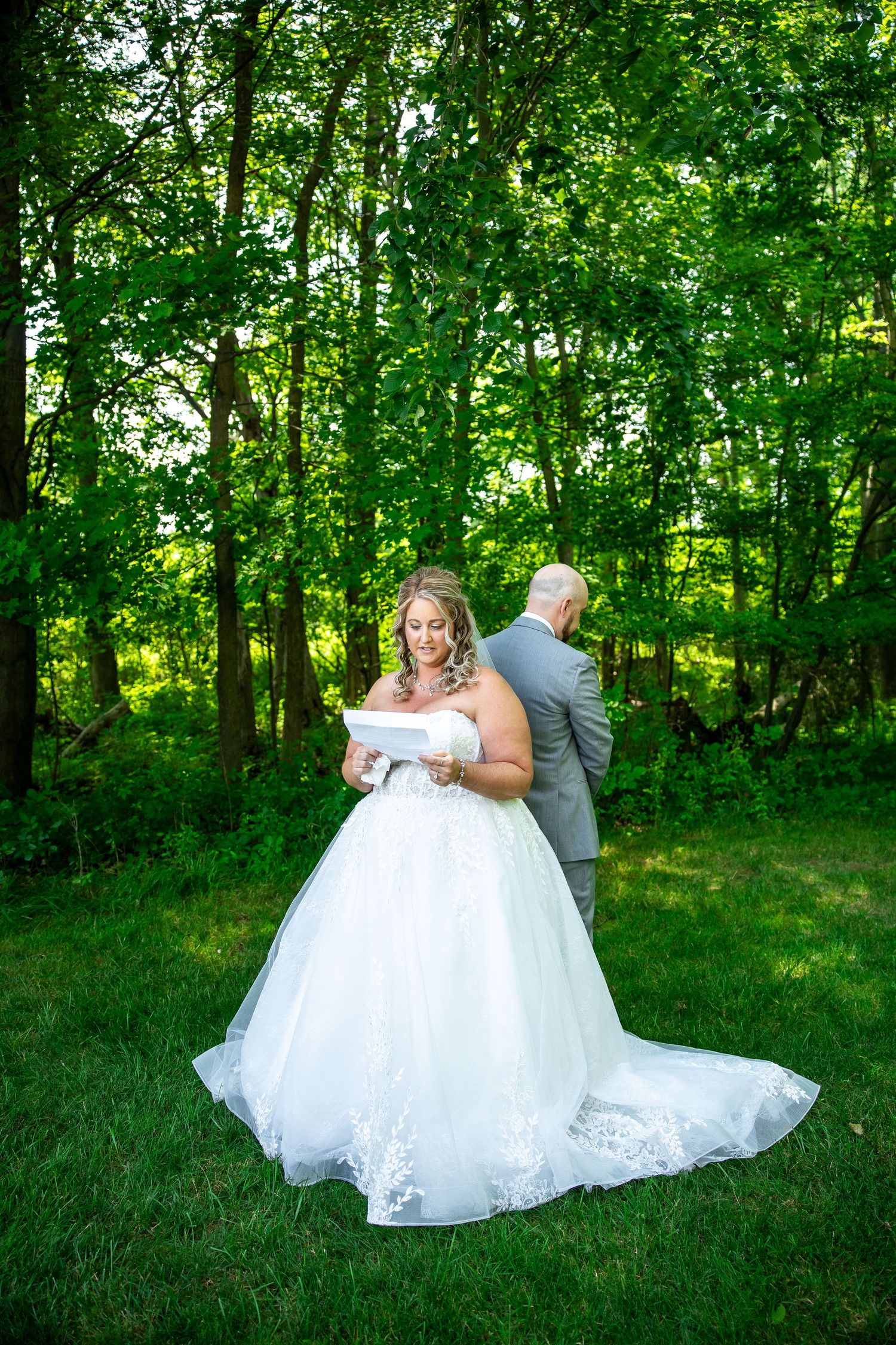 Bride in a white lace ball gown reads a letter while the groom stands with his back to her in a sunny, green wooded setting.
