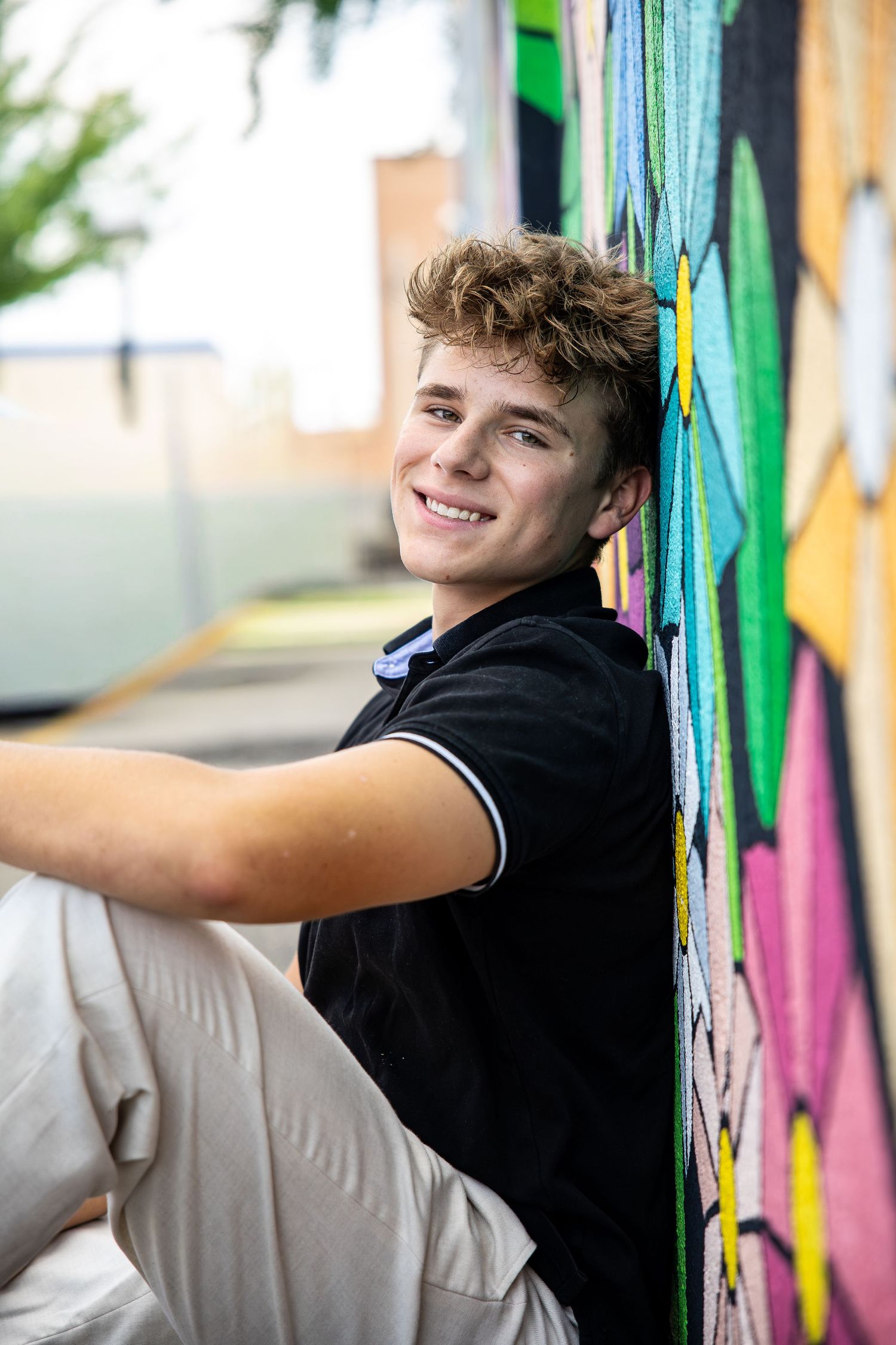 A smiling young man in a black polo shirt sits against a colorful graffiti wall outdoors.