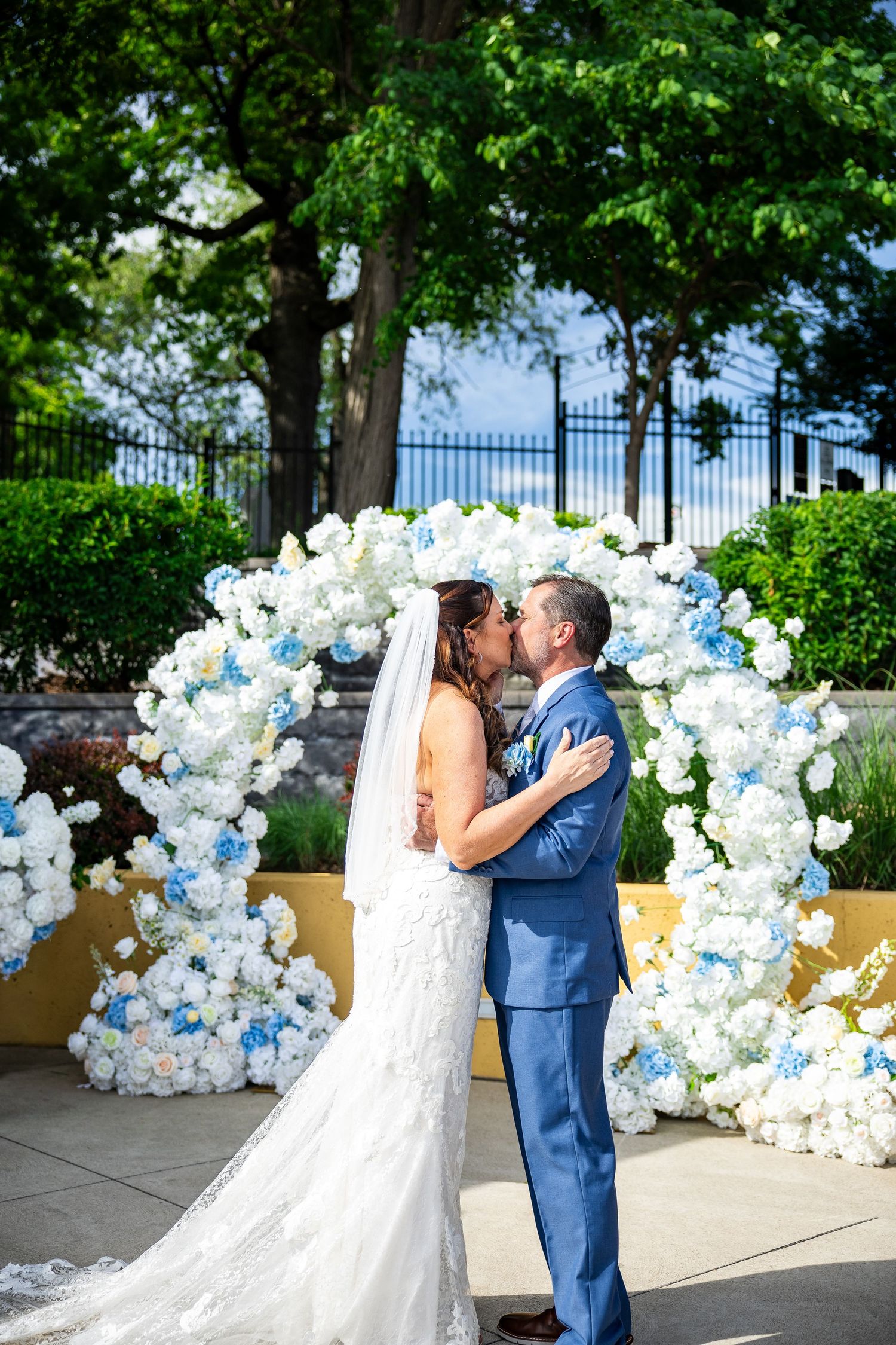 Bride in a lace gown and groom in a blue suit kiss beneath a white and blue floral wedding arch in a garden.