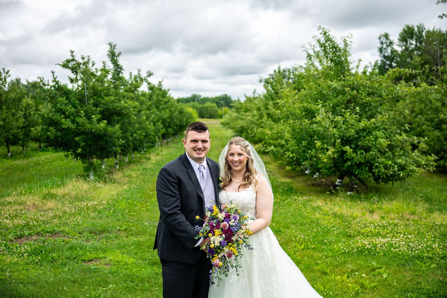 Bride and groom posing in a lush green orchard, bride holding a vibrant wildflower bouquet on their wedding day.