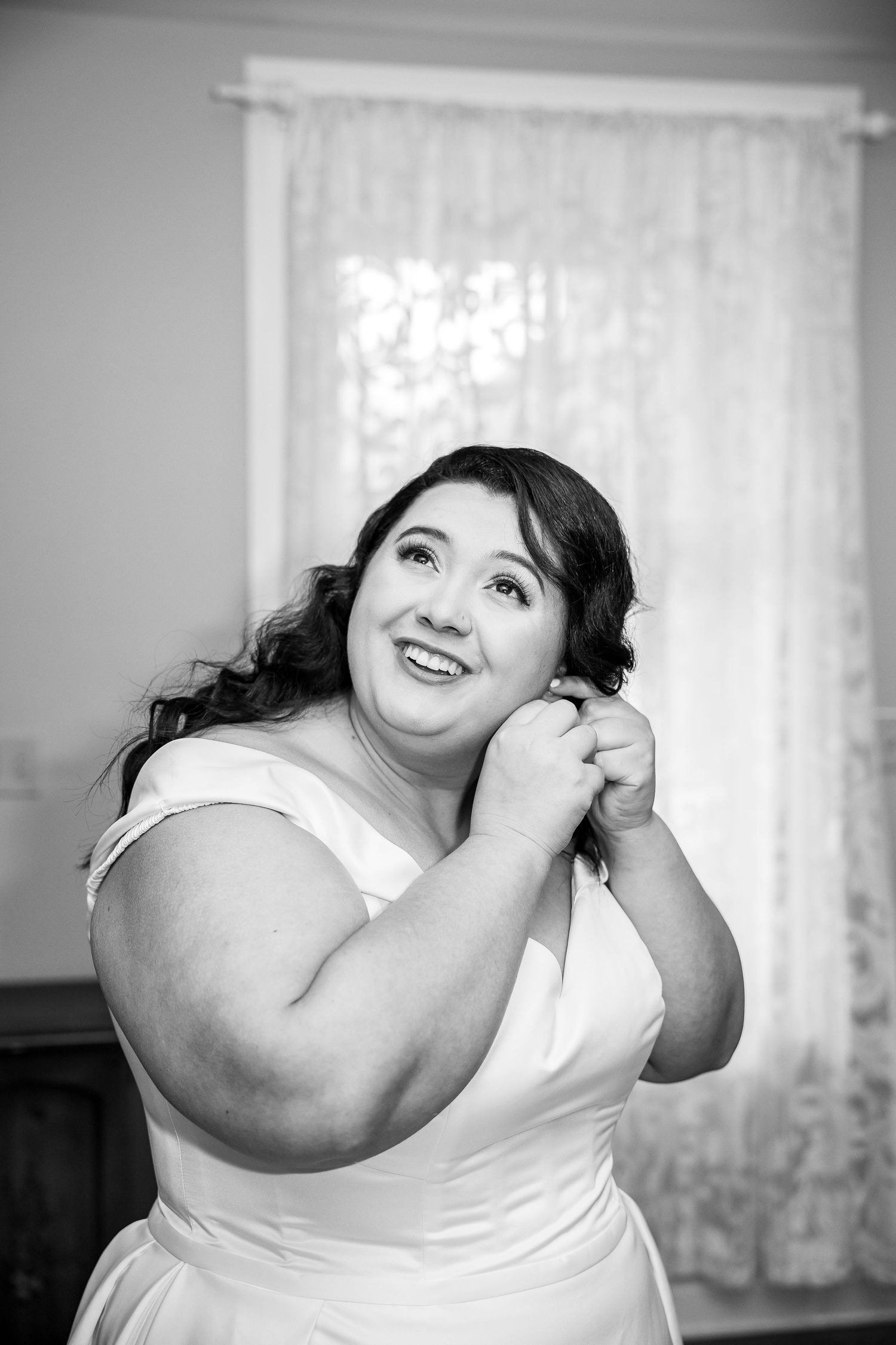 Black and white photo of a smiling bride in a white dress putting on earrings near a curtained window.