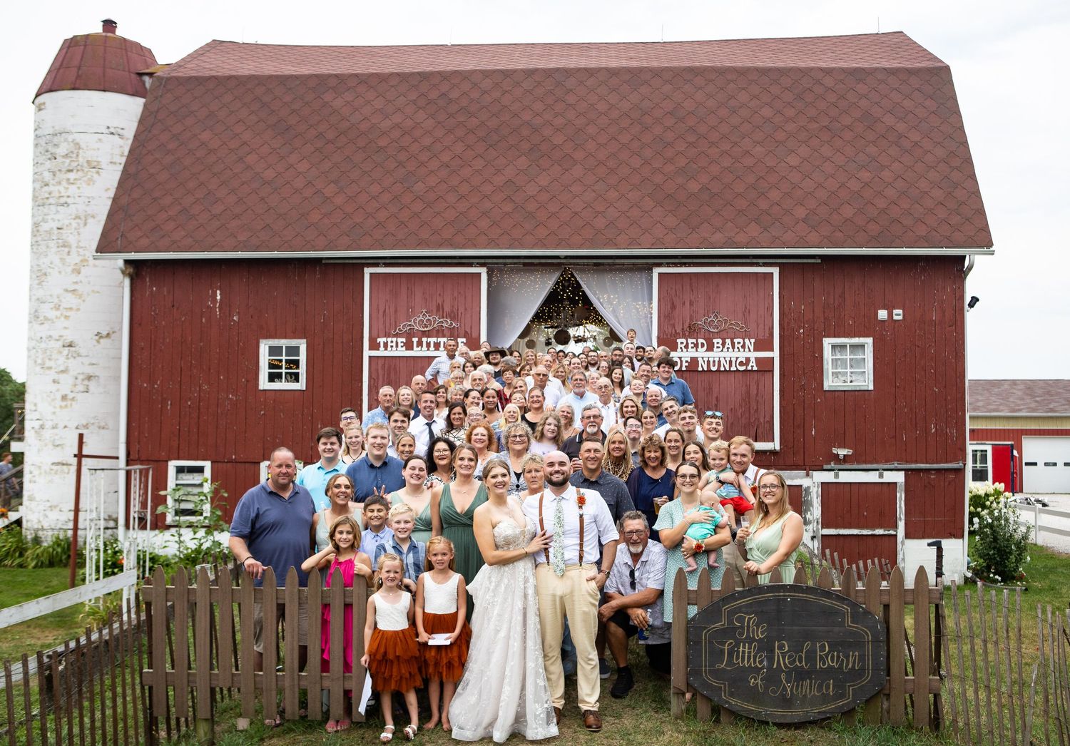 Large wedding party poses in front of a rustic red barn venue called The Little Red Barn on a sunny day.