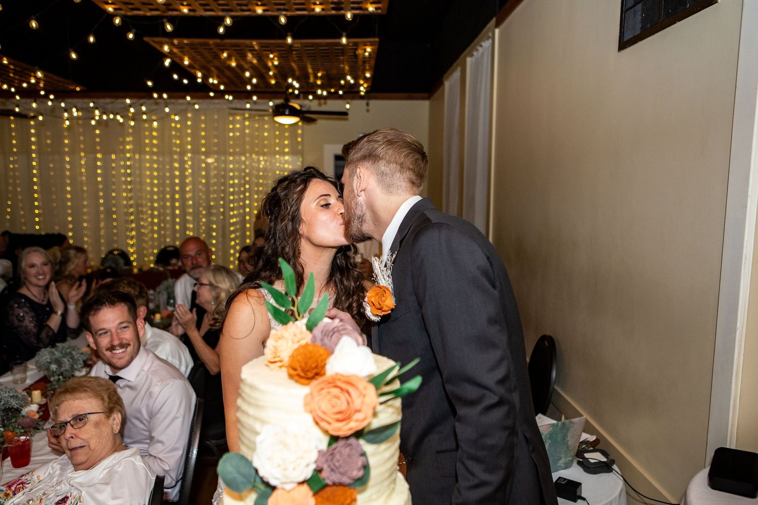 Bride and groom share a kiss while cutting their wedding cake decorated with orange and white flowers at their reception.