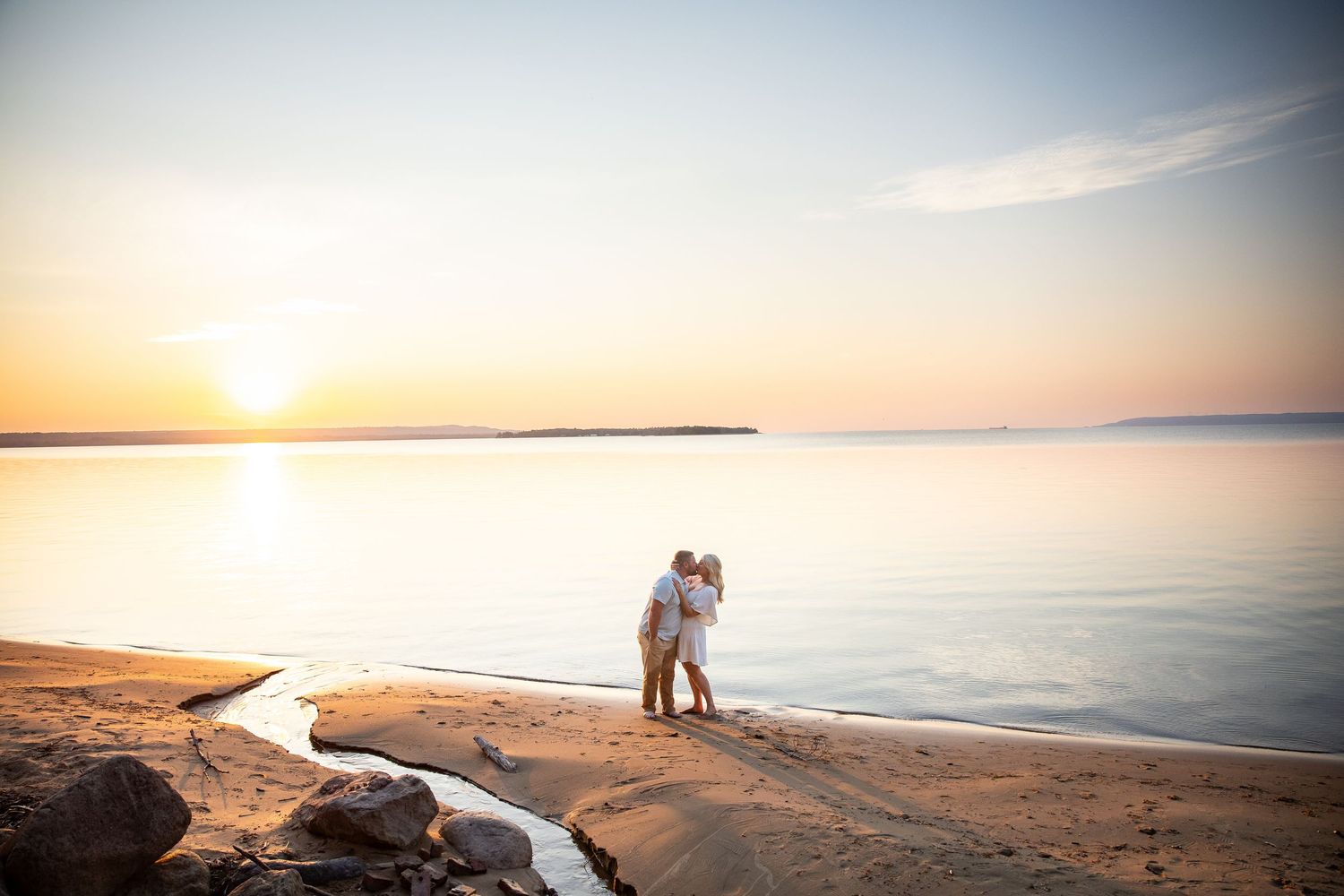 Couple embracing on sandy beach at golden sunset with calm water and soft pastel sky in background.