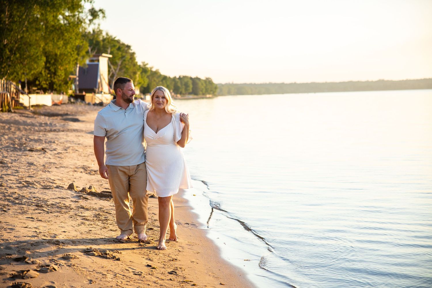 Couple walking hand in hand along a sandy beach at golden hour near calm water with trees in the background.