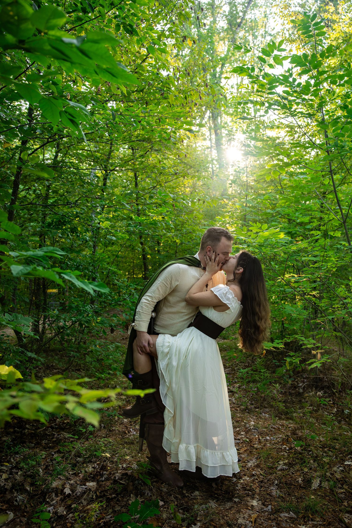 Couple sharing a romantic kiss in a sunlit forest, man lifting woman in white dress among lush green trees.