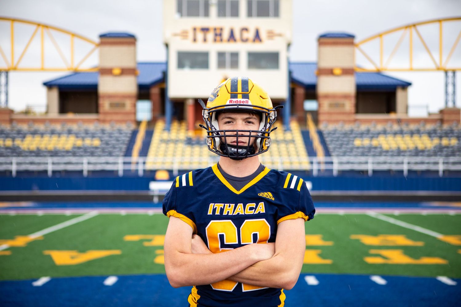 Football player in blue and yellow uniform stands with arms crossed on stadium field during golden hour.