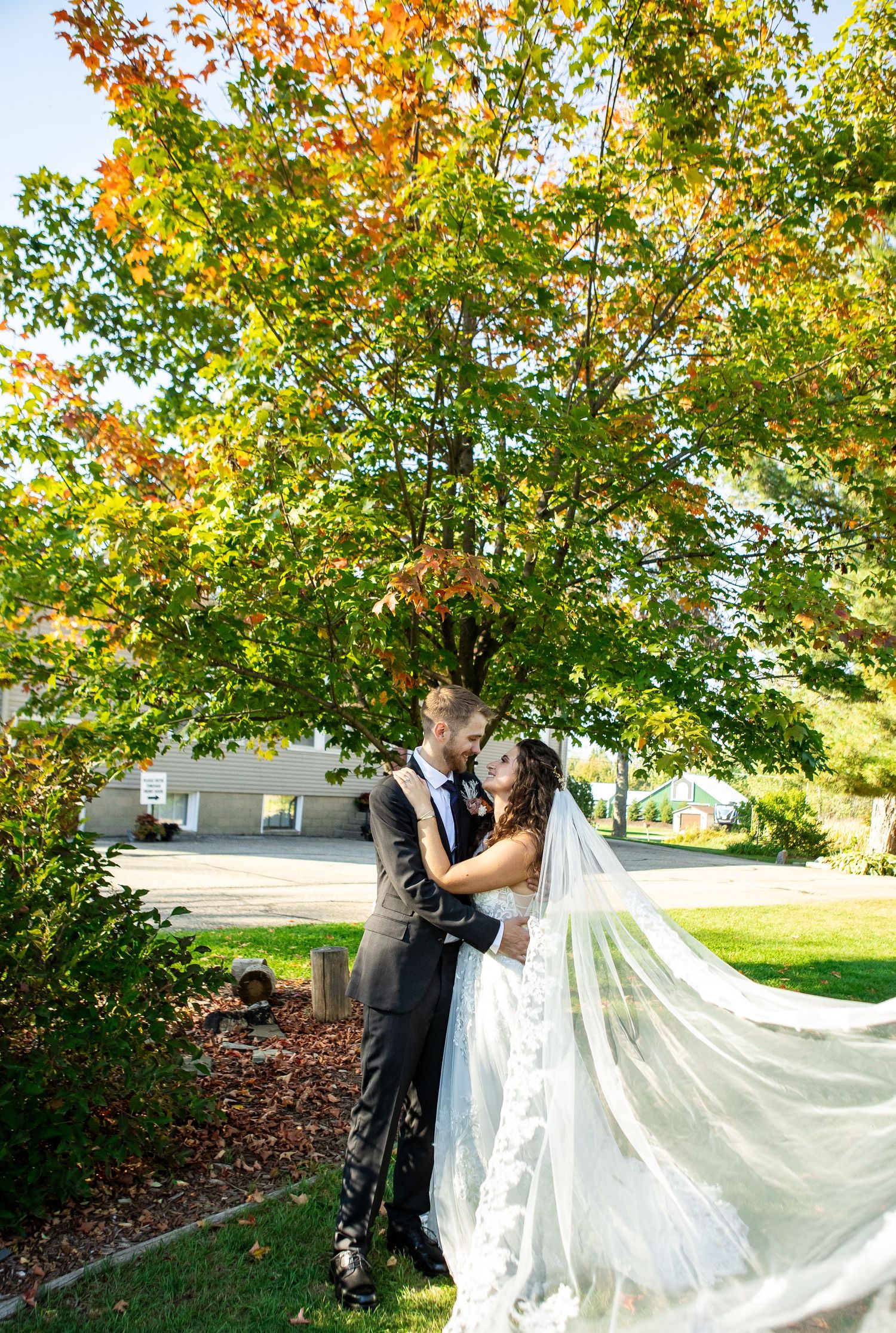 Bride and groom share a romantic moment under a colorful autumn tree, with her veil flowing gracefully in the breeze.
