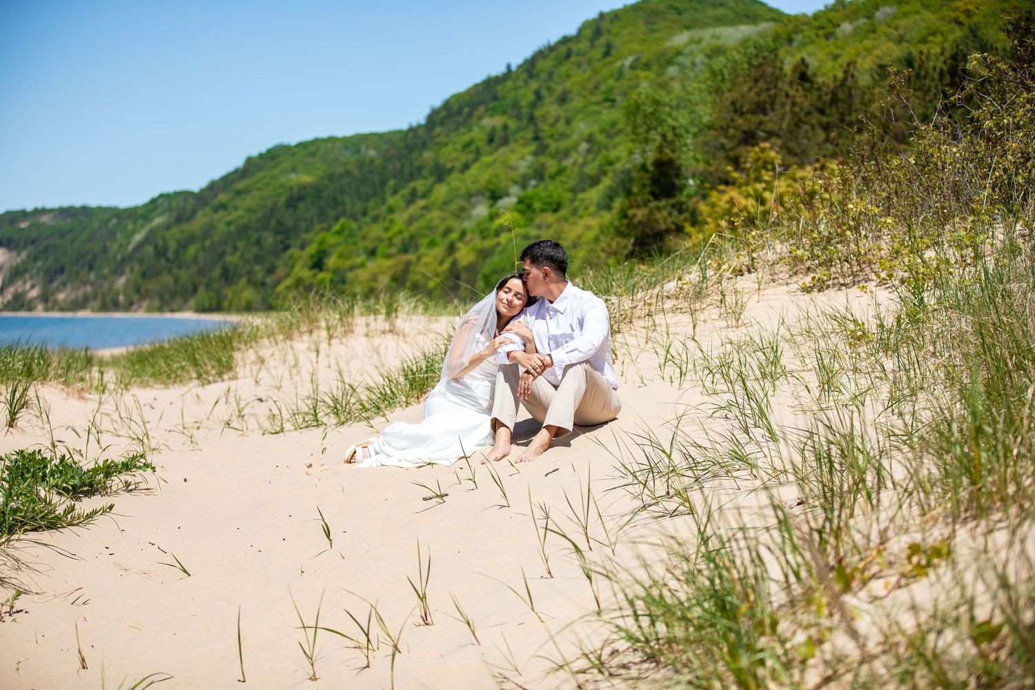 Couple in white sitting close together on a sandy dune with beach grass, overlooking a lake and green forested hills on a sunny day.