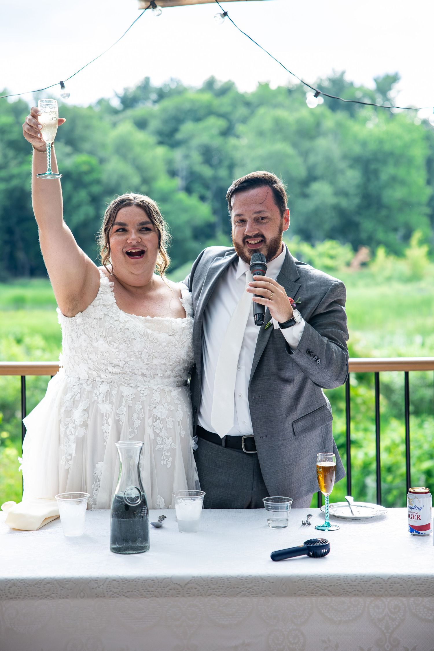 Bride raises champagne glass while groom speaks into microphone at outdoor wedding reception with lush green backdrop.