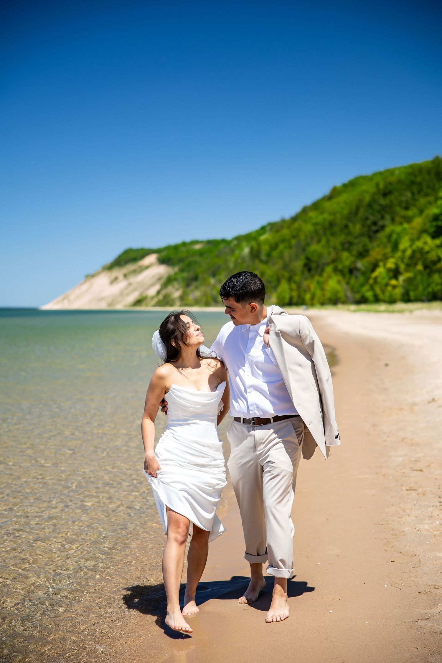 Newlywed couple walking barefoot on a sandy beach shore, bride in white dress, groom in light suit, green hills backdrop.