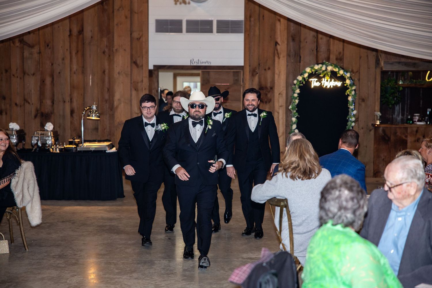 Groomsmen in black tuxedos make a grand entrance at a rustic barn wedding reception with wood-paneled walls.