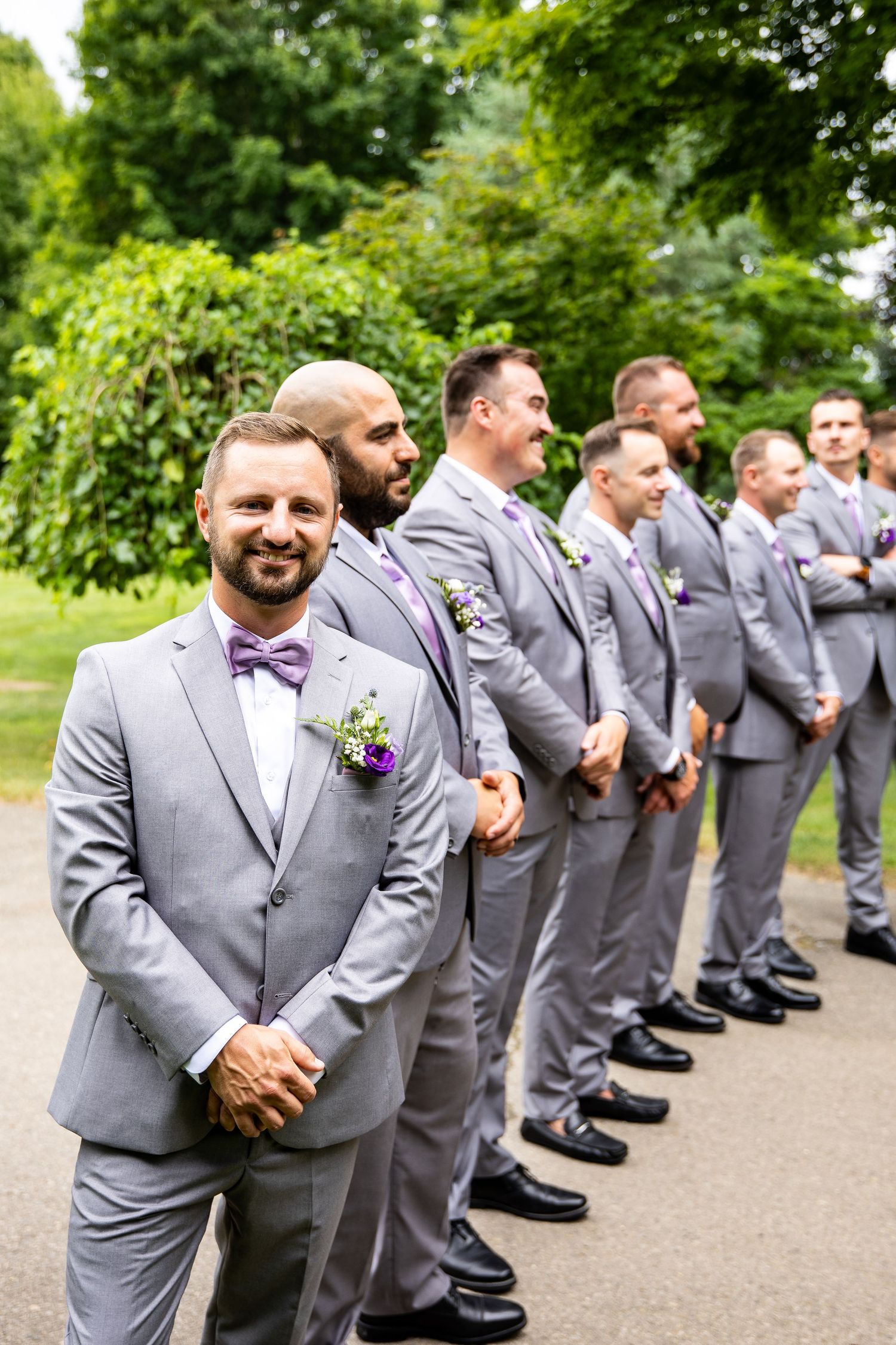 Groom in gray suit with purple bow tie stands before groomsmen in matching gray suits with purple boutonnieres outdoors.