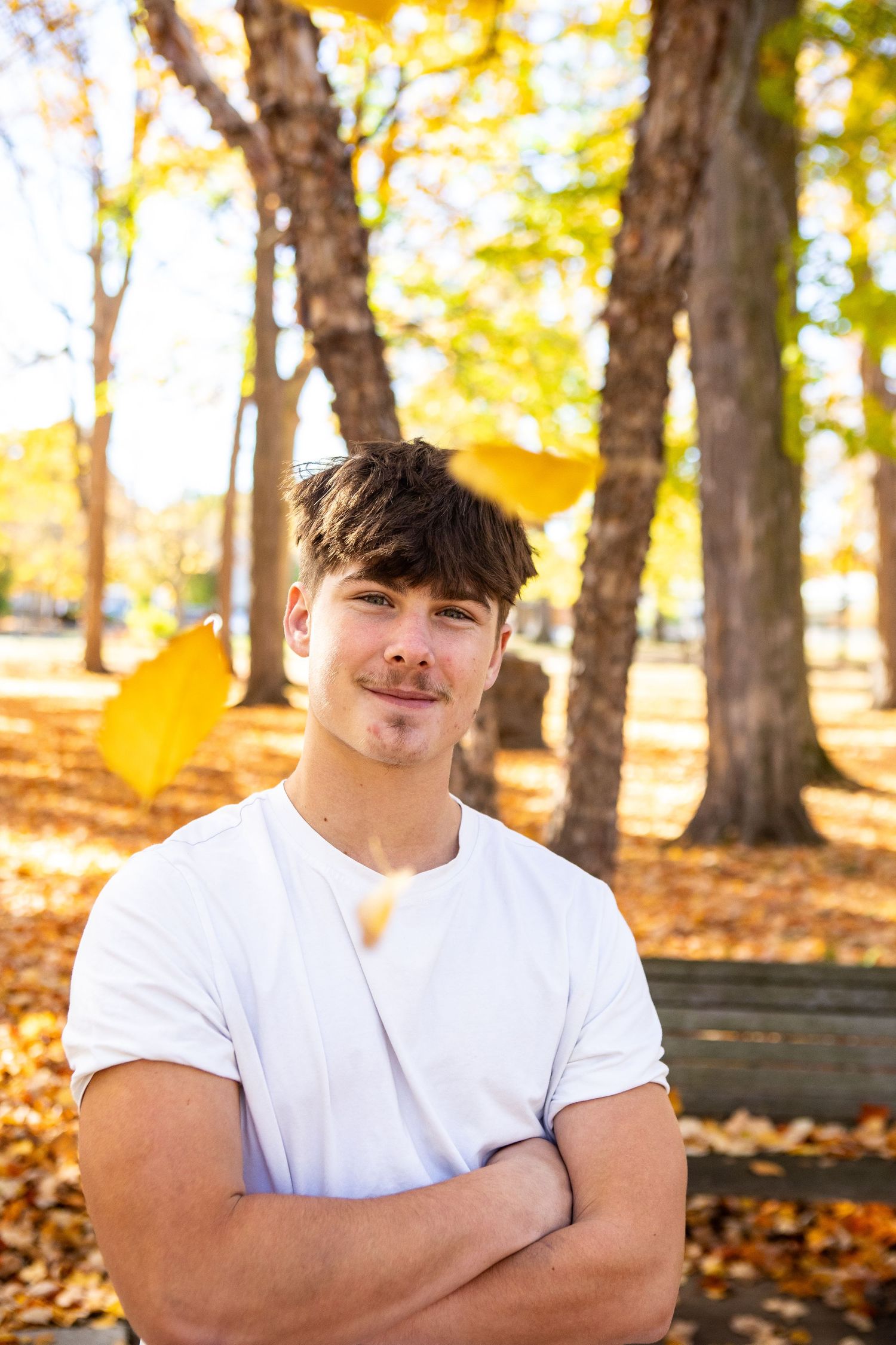 Young person in white t-shirt poses outdoors with autumn trees and golden leaves in background.
