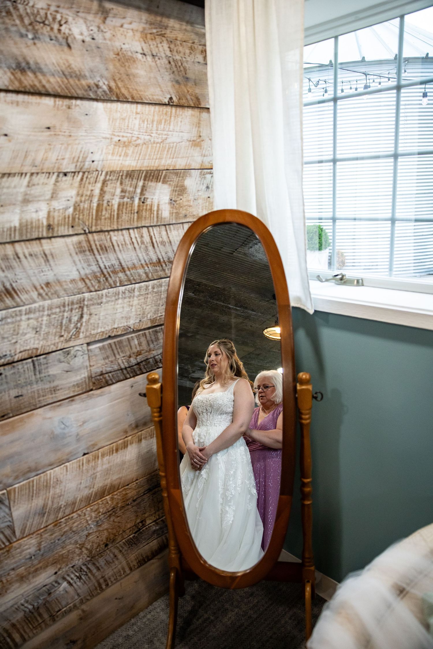 Bride in a white lace wedding gown and an older woman in purple are reflected in a wooden standing mirror beside a rustic plank wall.