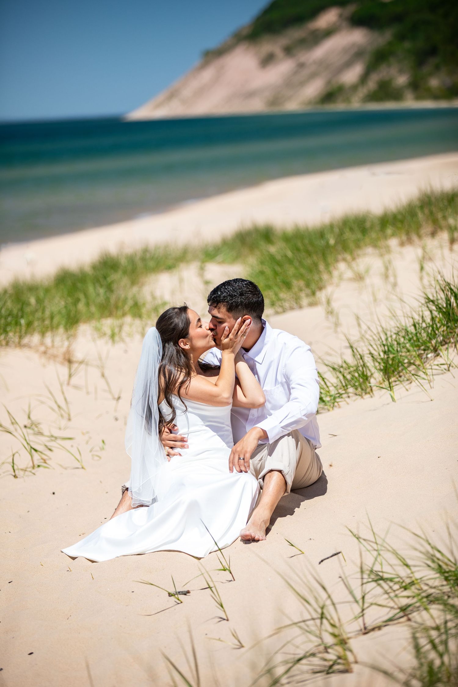 A bride and groom share a romantic kiss while sitting on sandy beach dunes with ocean and cliffs in the background.