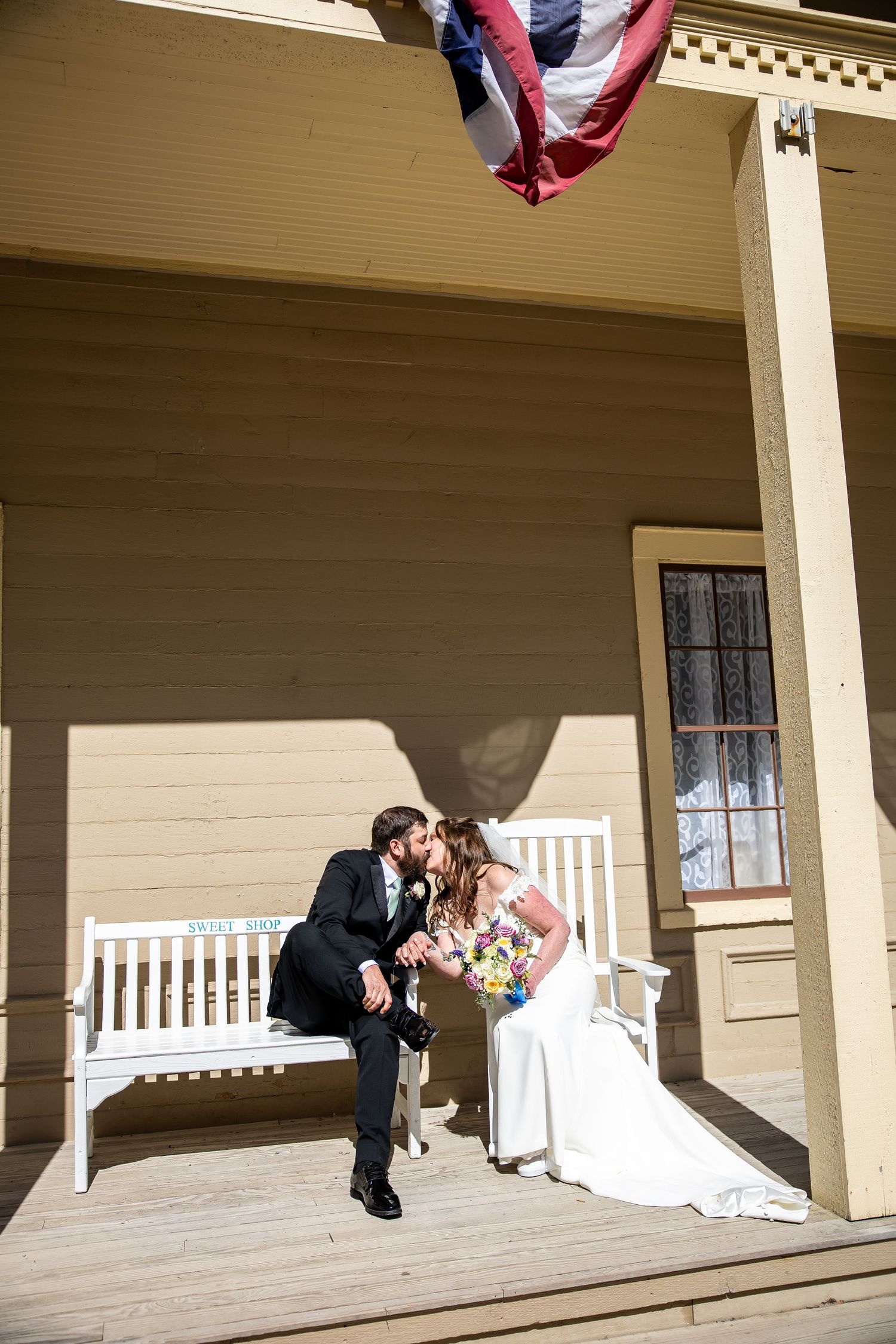 Bride and groom sharing a kiss on a sunlit wooden porch with a white bench and American flag backdrop.