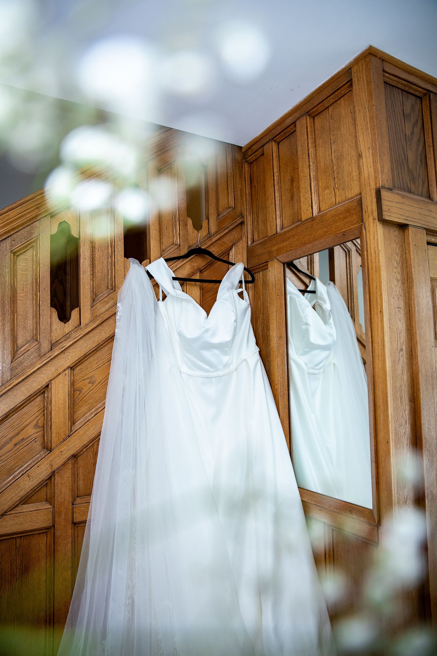 White wedding dress with veil hanging on wooden wardrobe, reflected in mirror, with soft floral bokeh foreground.