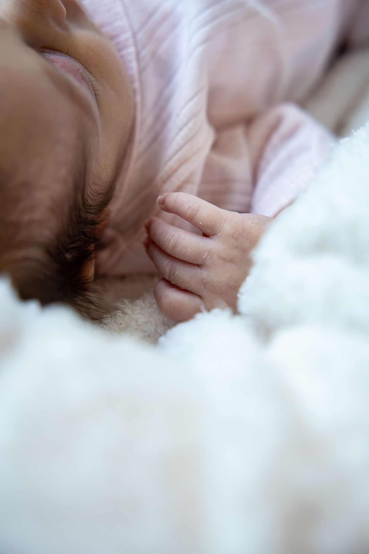 Close-up of a sleeping newborn baby in pink outfit with tiny curled fingers resting on soft white fluffy blanket.