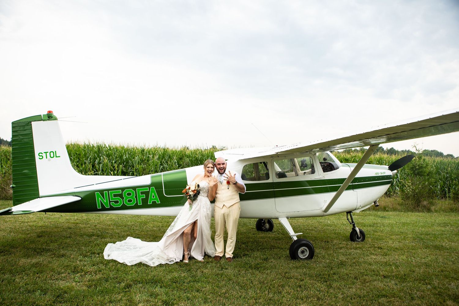 Bride and groom pose beside a green and white Cessna plane on a grass airfield on their wedding day.