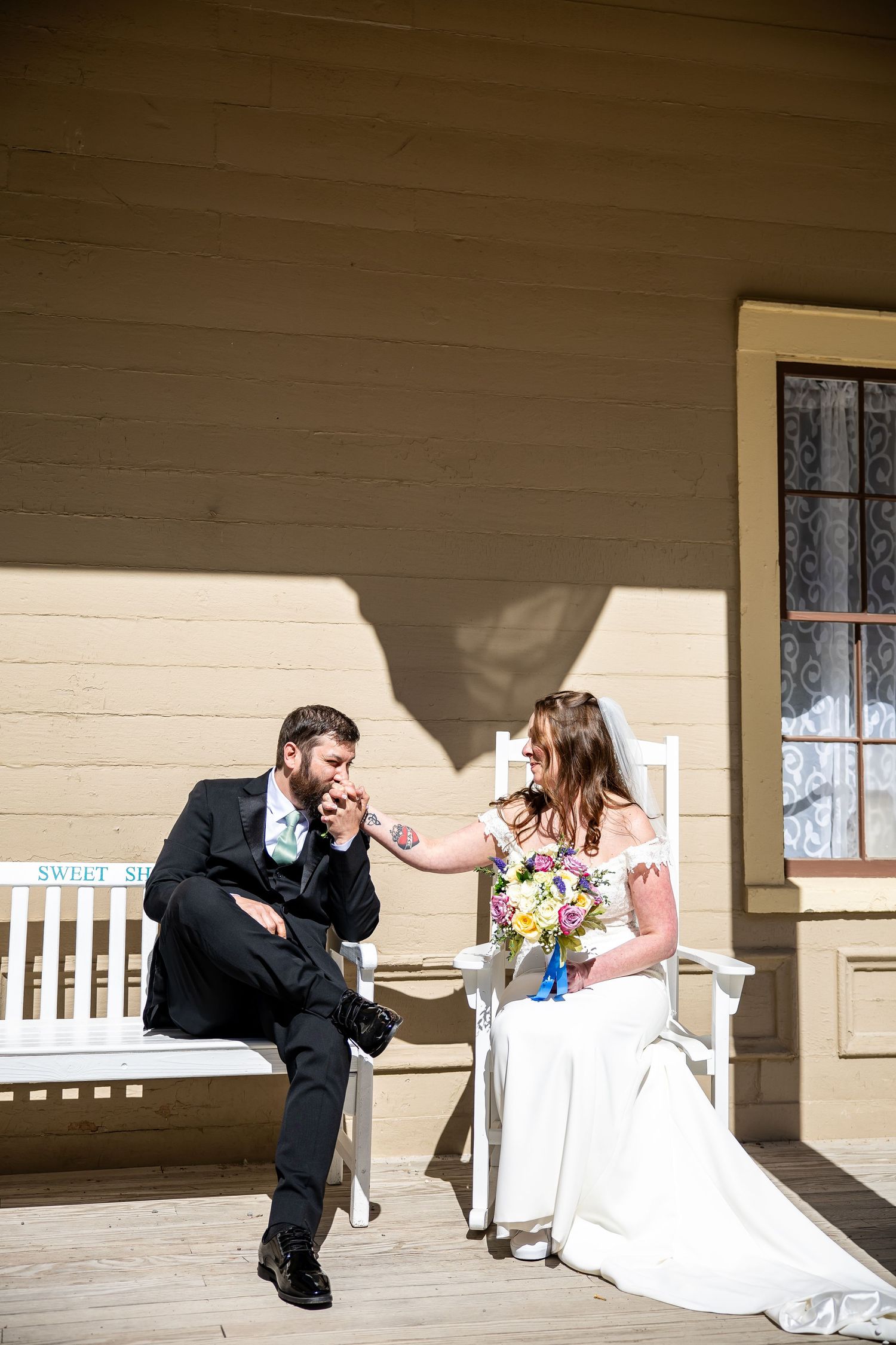 Bride and groom sitting on white chairs on a sunny porch, sharing a tender moment with a colorful bridal bouquet.