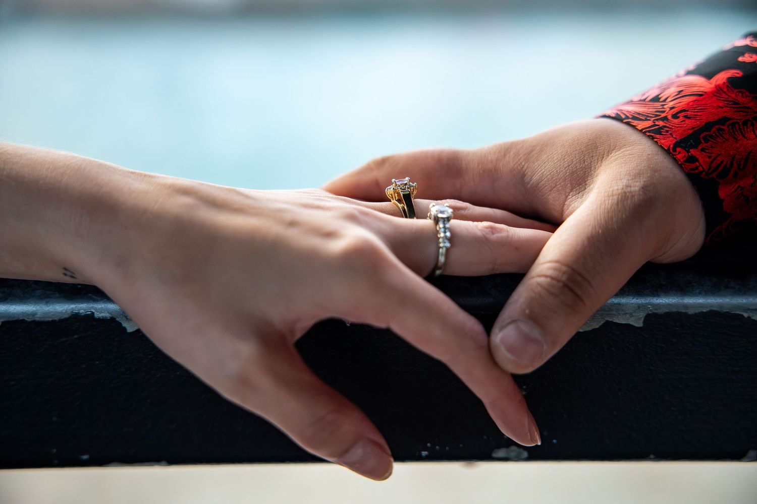 Two hands touching on a railing, one wearing a diamond engagement ring and a gold ring, with water in the background.