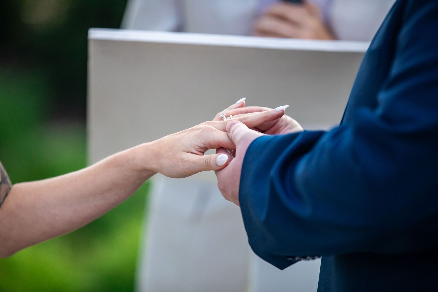 Bride and groom exchanging rings during an outdoor wedding ceremony as an officiant stands in the background.