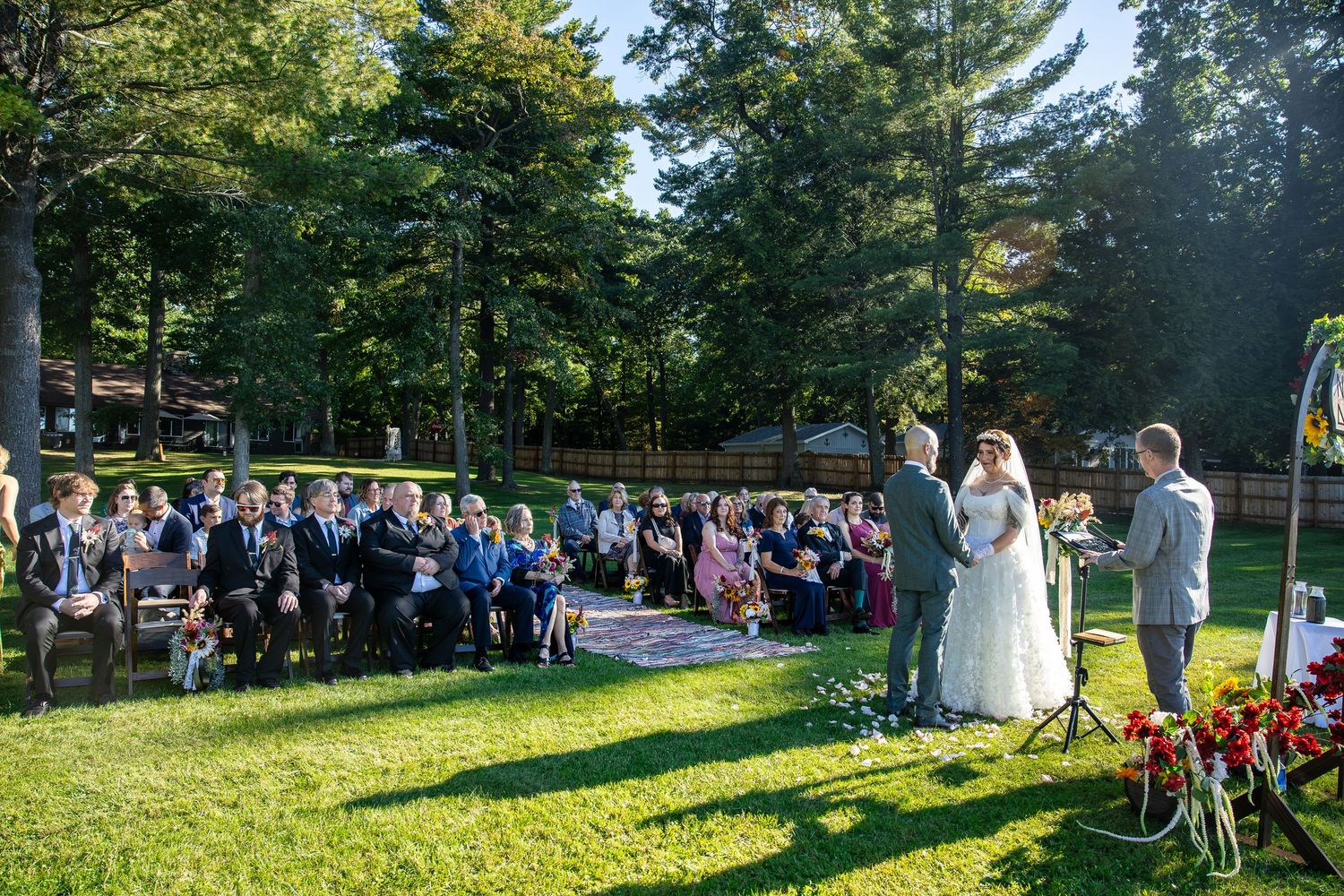 Bride and groom exchange vows before seated guests during an outdoor wedding ceremony on a sunlit lawn surrounded by tall evergreen trees.