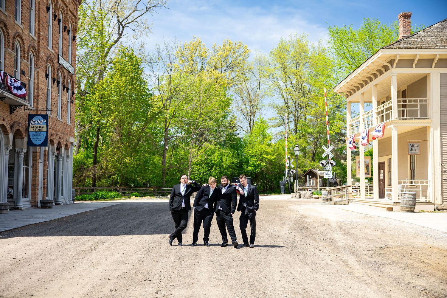 Four groomsmen in black suits walking together on a sunny dirt road at a historic outdoor venue.