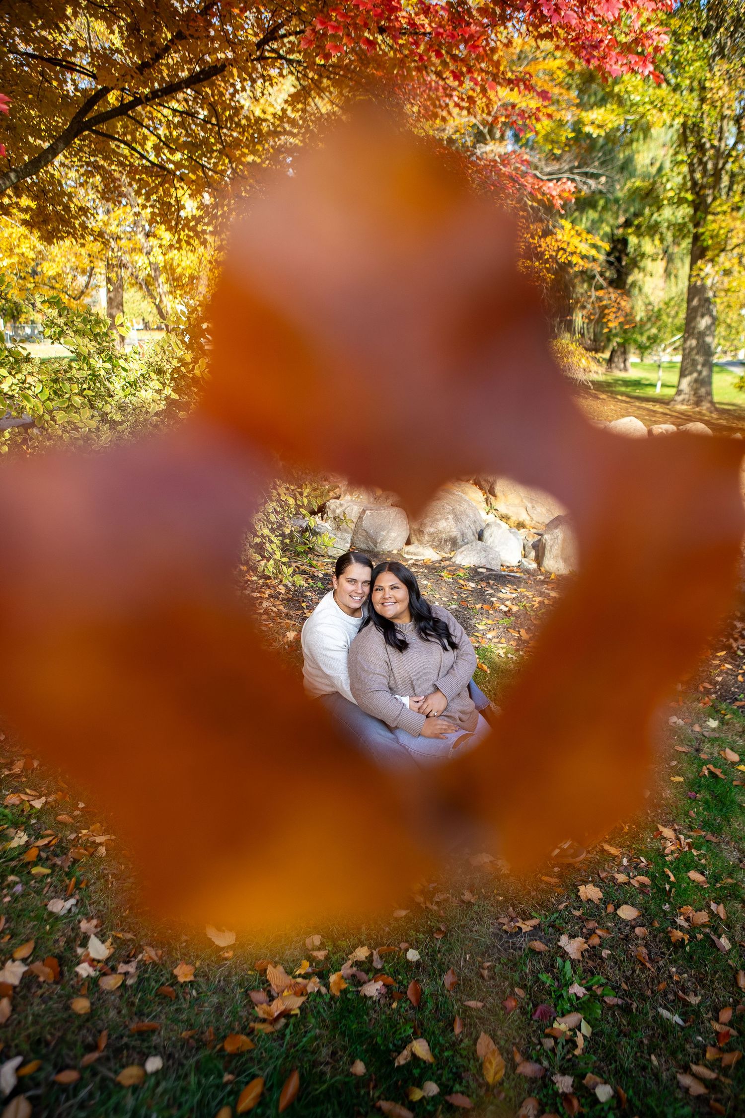 Couple embracing in autumn park, viewed through heart-shaped frame of vibrant orange and red fall leaves.