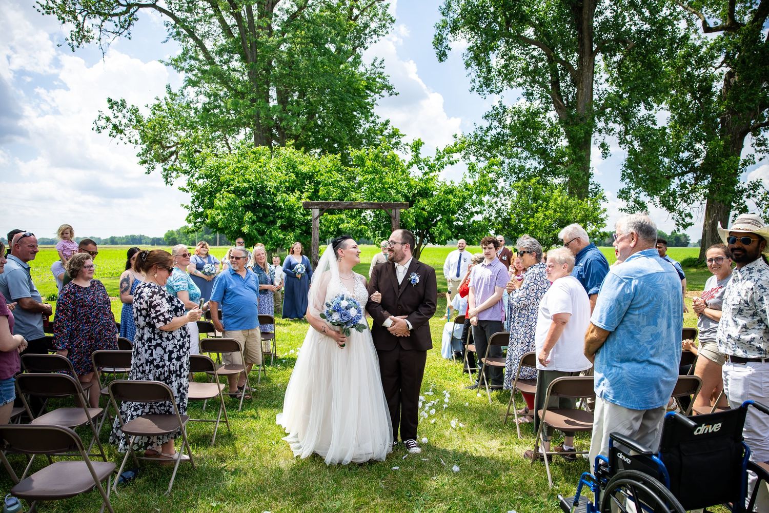 Bride and groom walk down outdoor wedding aisle surrounded by guests on a sunny day with lush green trees.