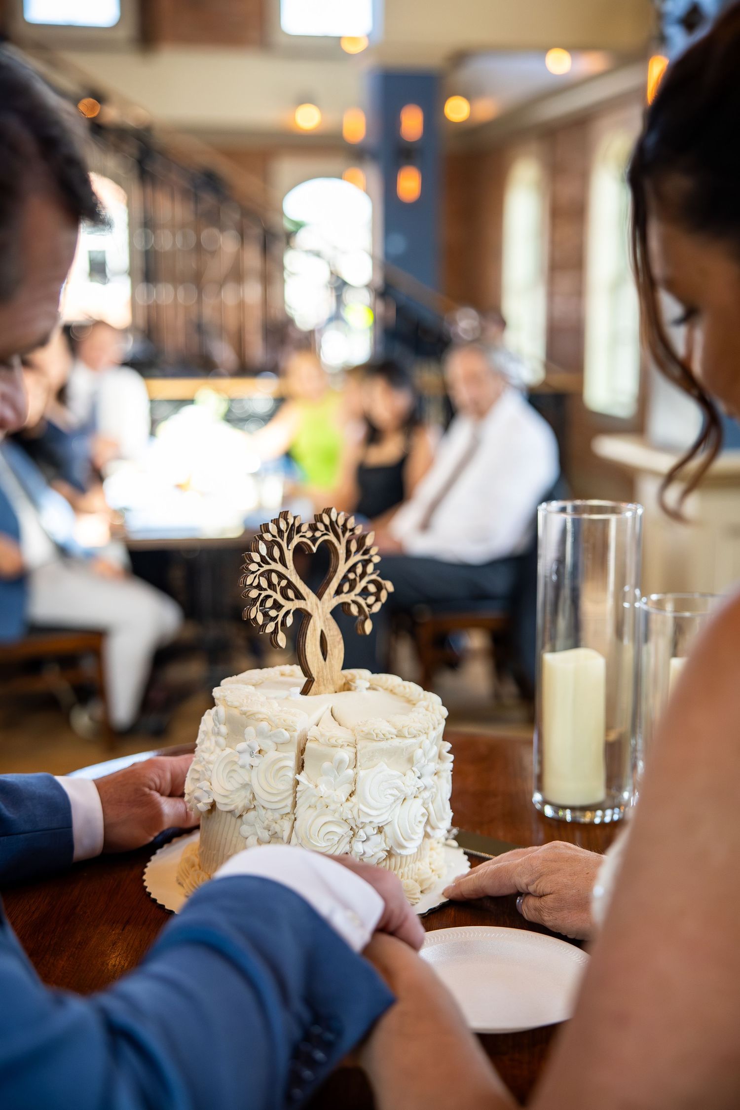 Couple cutting a white wedding cake with a wooden tree heart topper in a warmly lit restaurant reception venue.