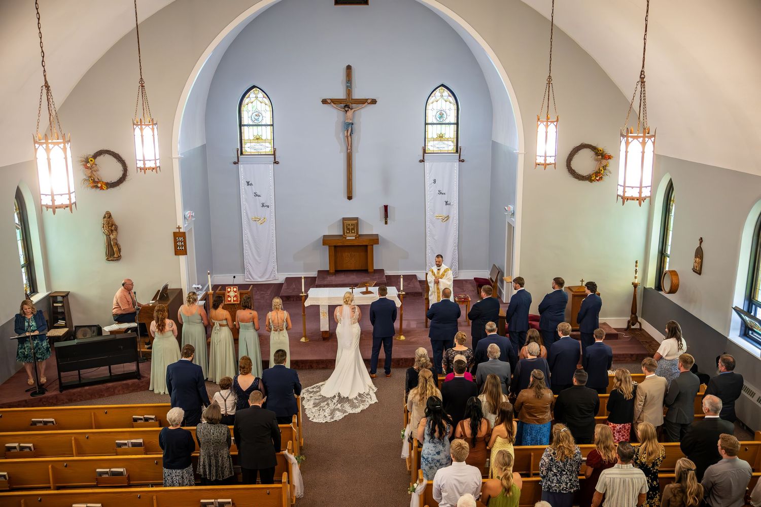 Bride in white gown and groom stand at the altar during a church wedding ceremony as bridesmaids, groomsmen, and seated guests look on.