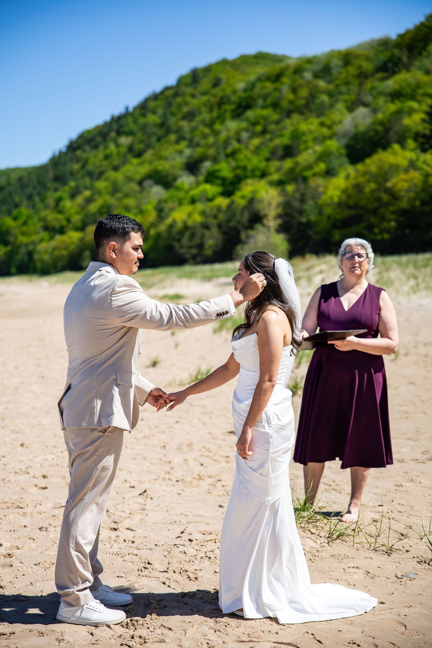 Couple exchanging vows on sandy beach with officiant in purple dress and lush green hills under blue sky.