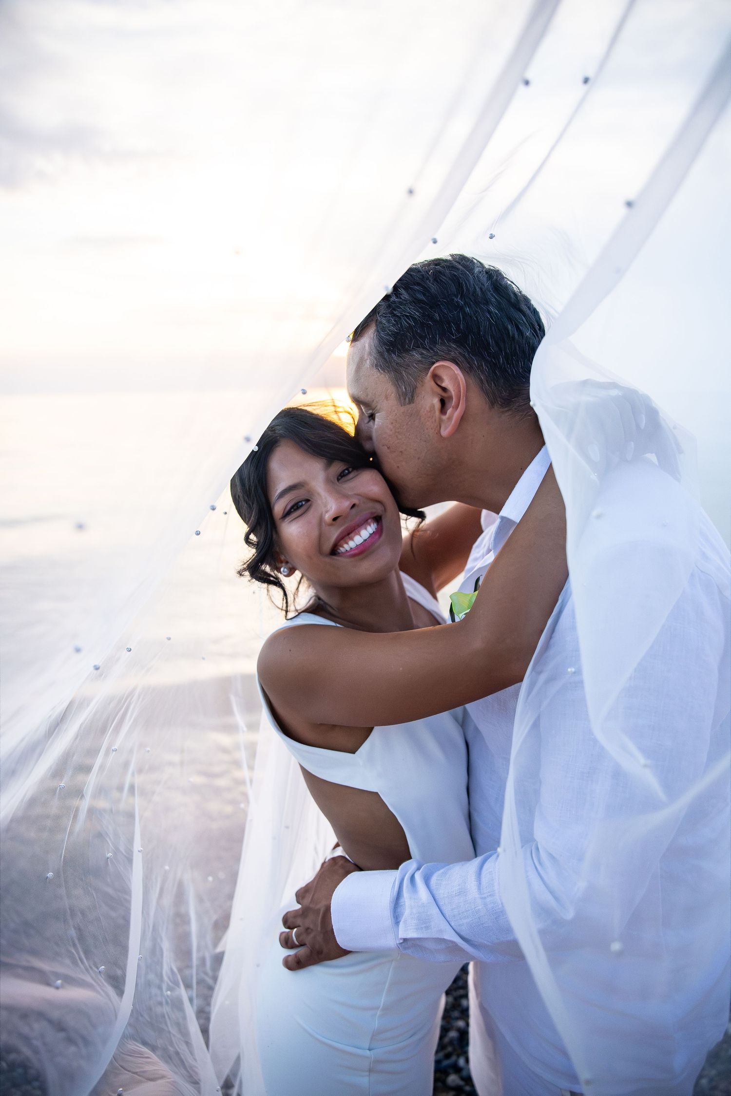 A smiling bride in a white dress is kissed on the cheek by her groom as a sheer veil billows around them on a beach at sunset.