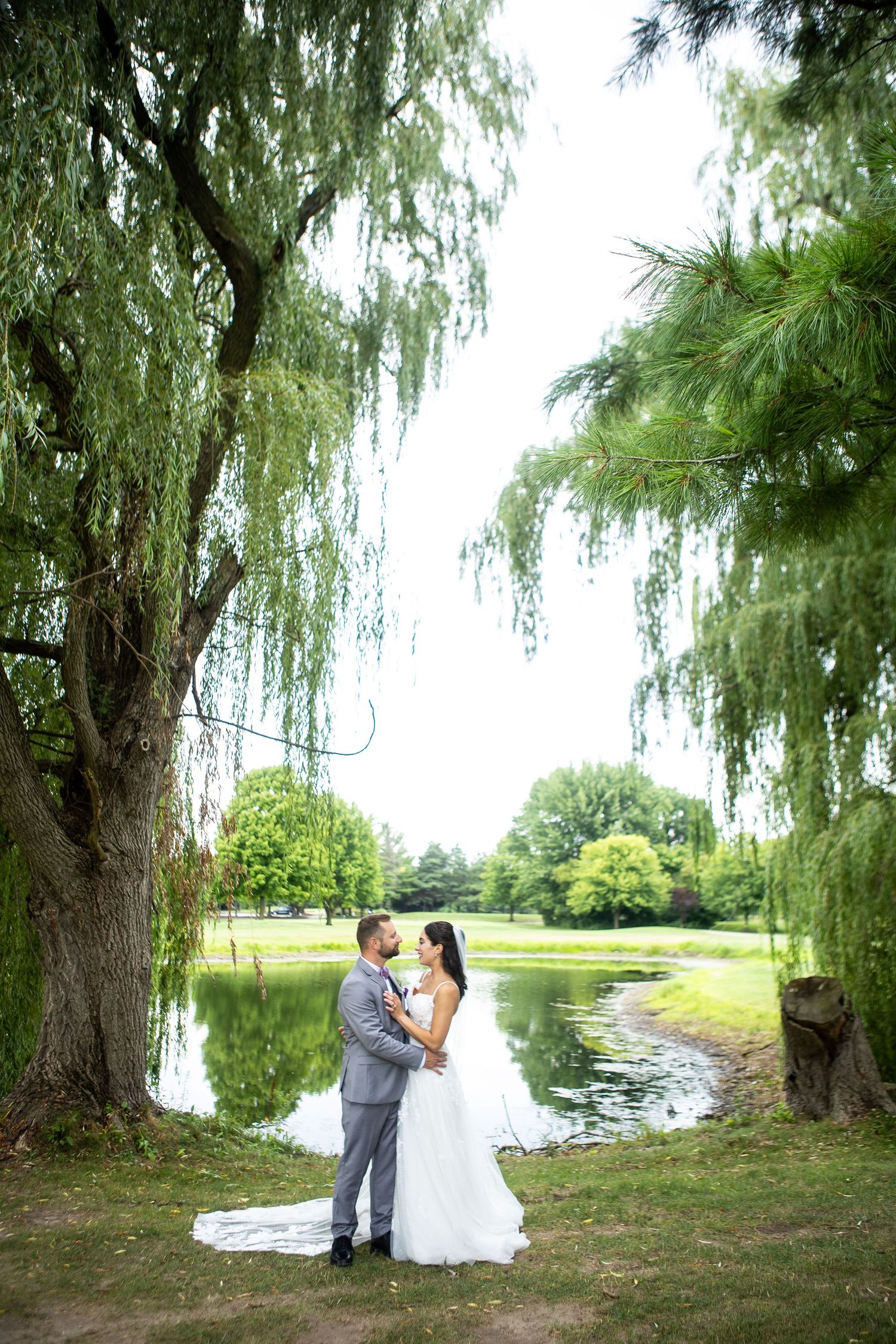 Bride and groom embrace under weeping willow tree by serene pond in lush green park on wedding day.