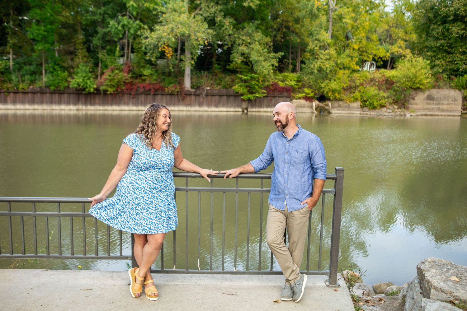 A couple holds hands while standing by a lake railing on a sunny day.