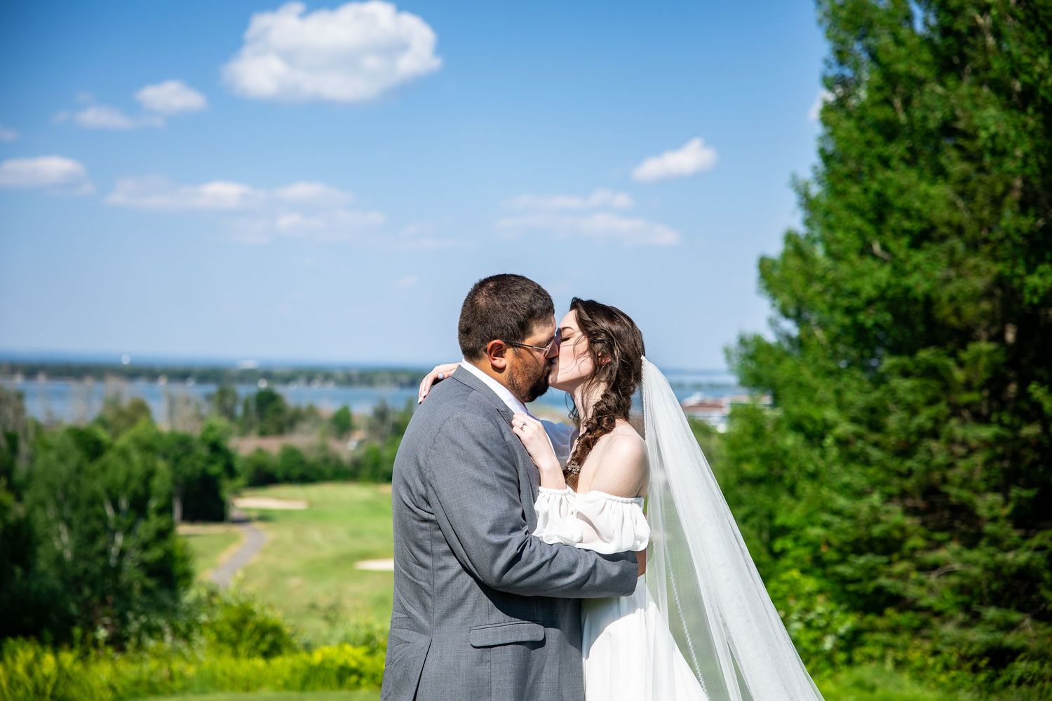 Bride in a white gown and veil kisses her groom in a gray suit on a sunny golf course overlooking a scenic waterfront and trees.