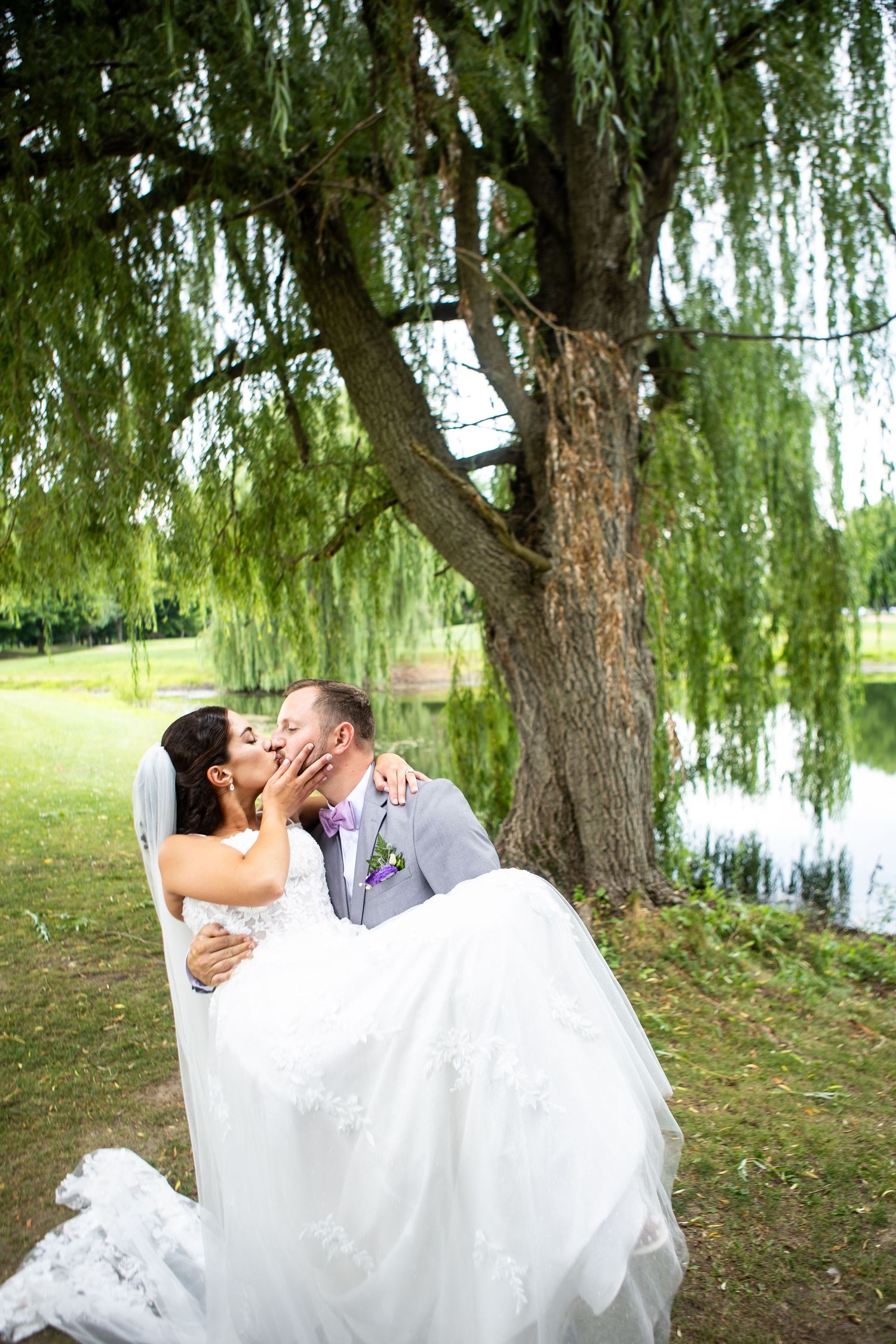 Groom carries bride in white lace gown, sharing a kiss under a weeping willow tree by a serene pond.