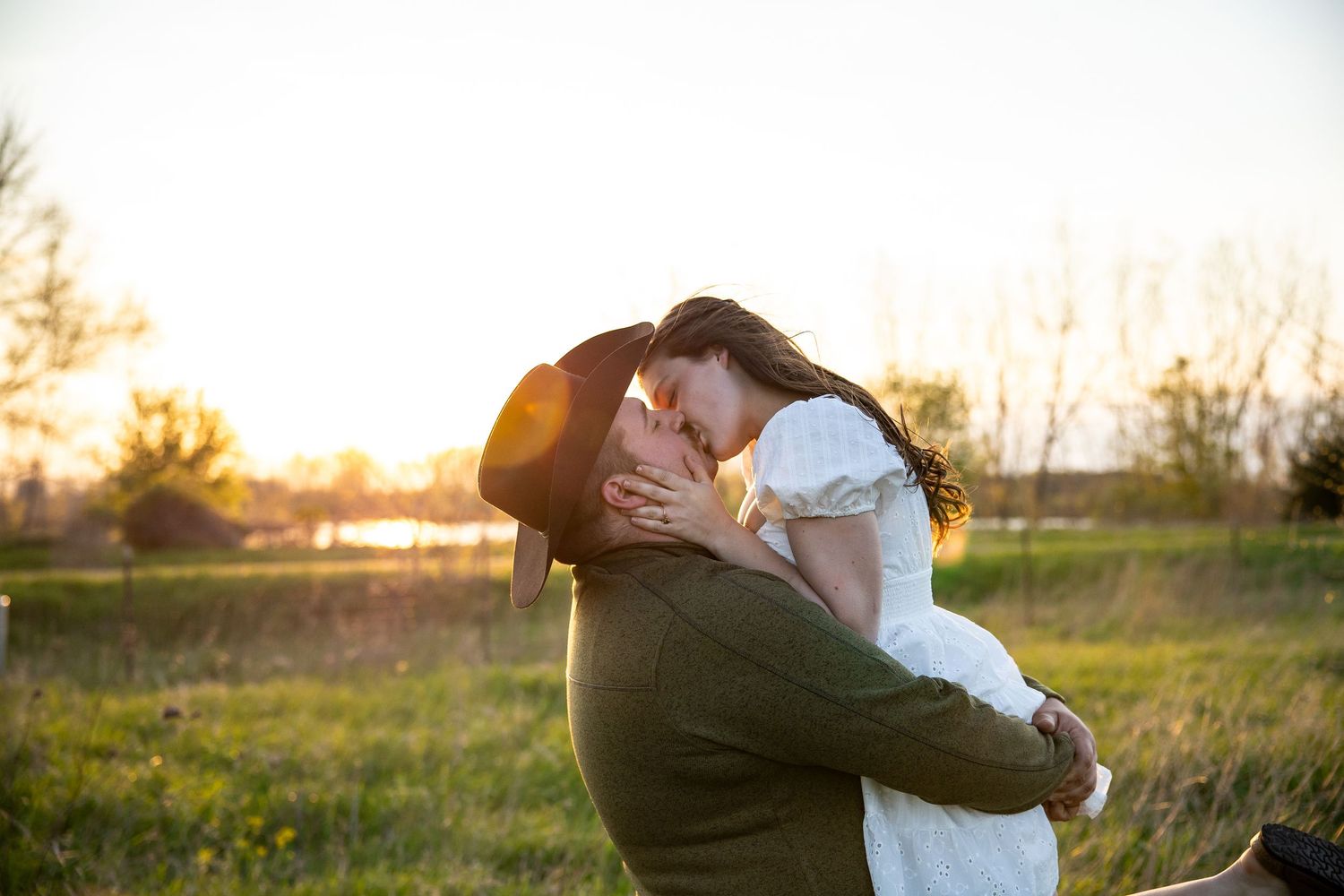 Couple sharing a romantic kiss at sunset in a green field, man lifting woman in white dress, golden hour lighting.
