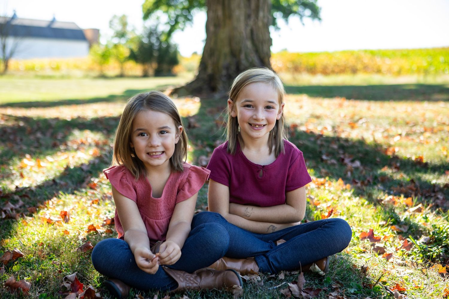 Two children sit cross-legged on autumn leaves in a sunny outdoor setting.