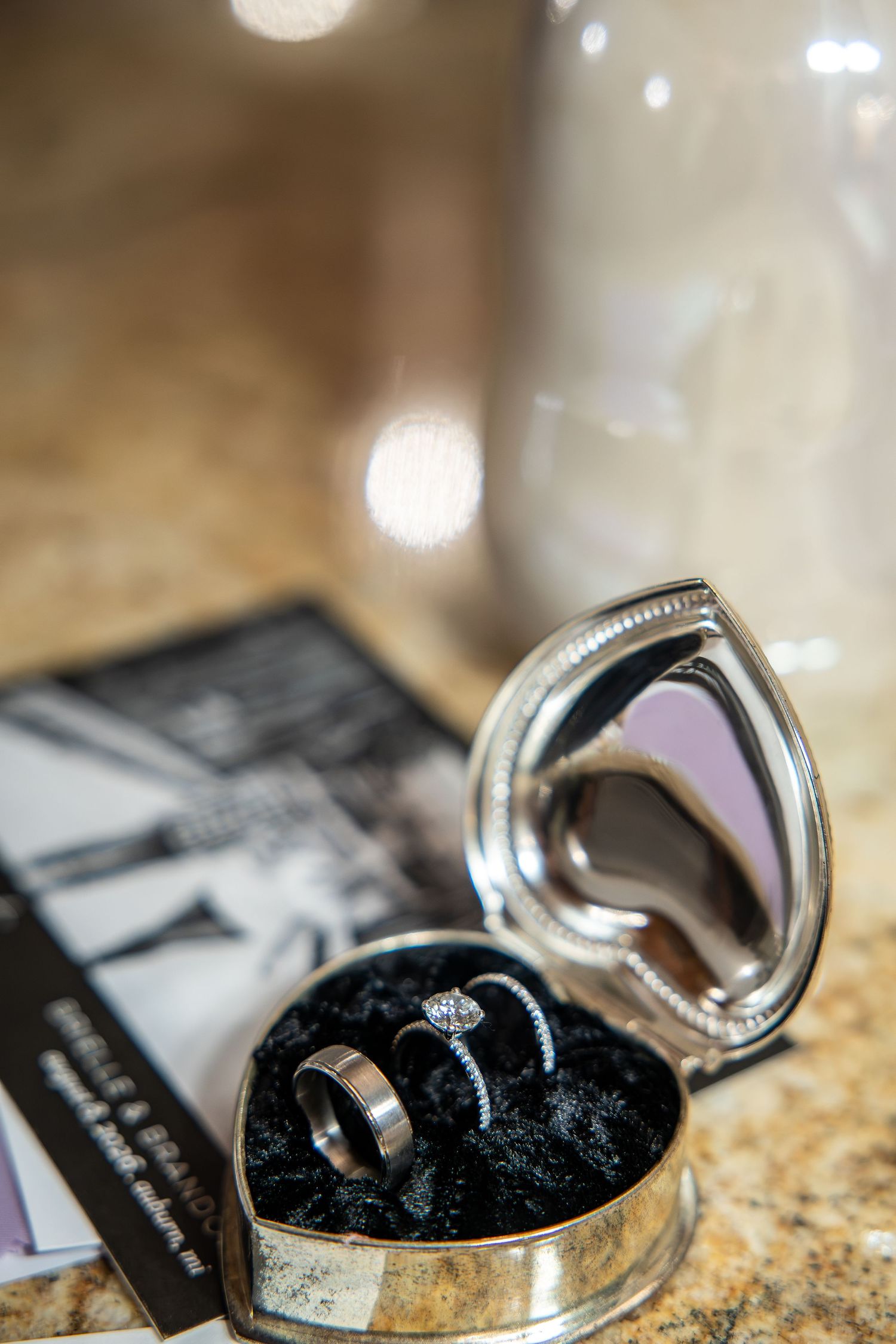Wedding rings and a diamond engagement ring rest on black velvet inside an open silver heart-shaped ring box with a mirror.