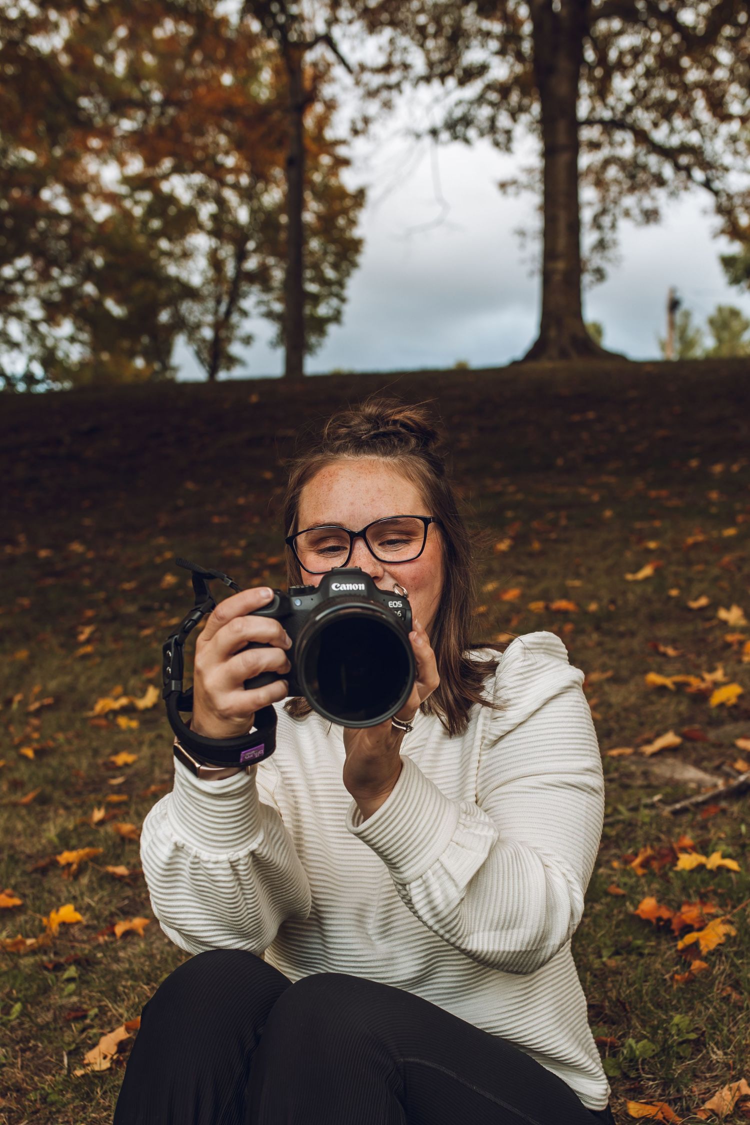 A photographer in a white sweater takes photos while lying on autumn leaves in a wooded setting.
