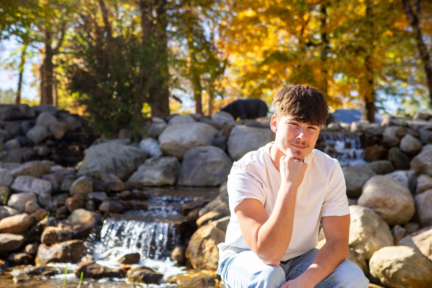 Someone in a white t-shirt sits thoughtfully by a stone waterfall on a sunny autumn day with golden foliage.