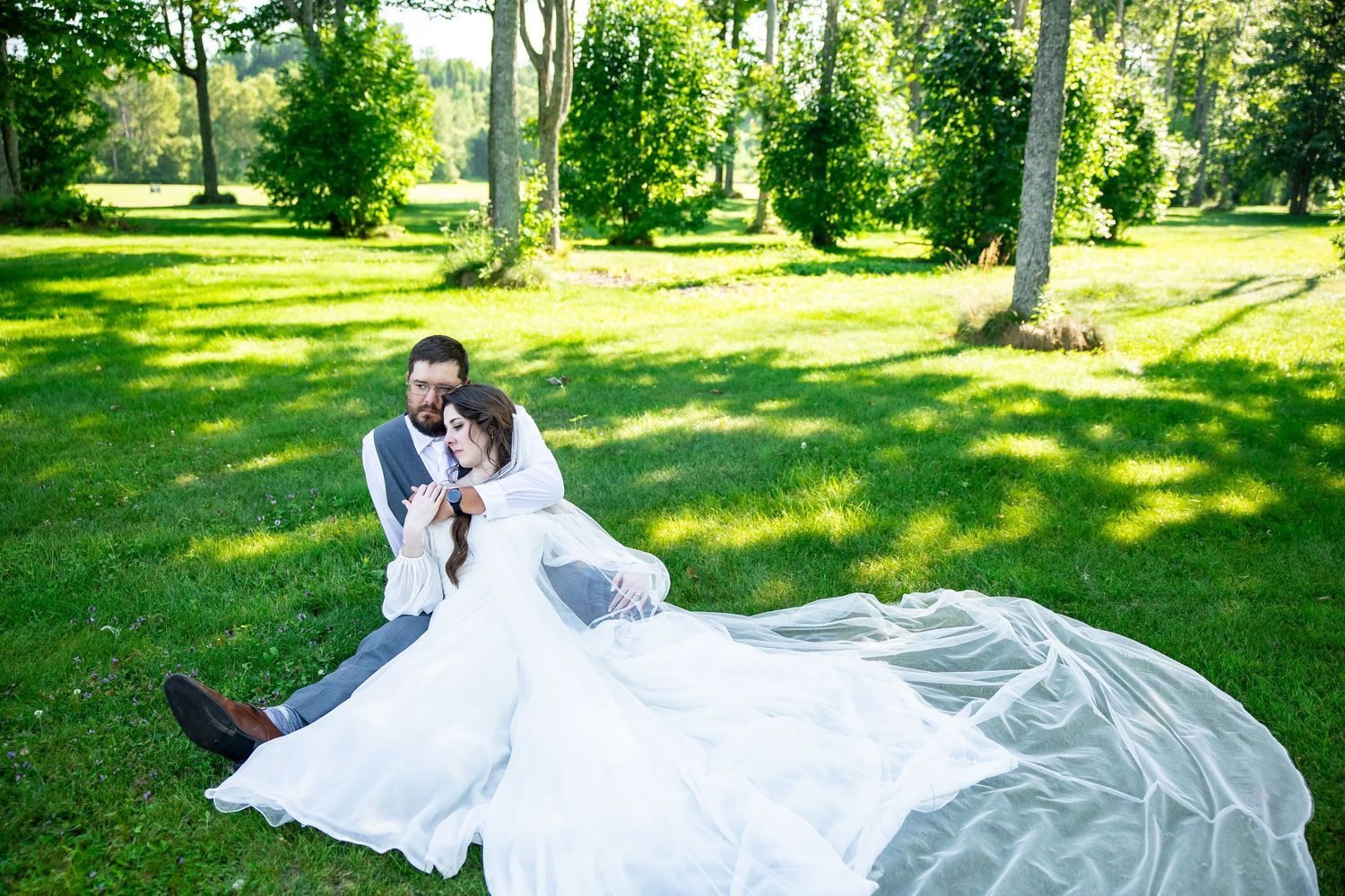 Bride and groom sitting on lush green grass in a park, bride's white veil flowing elegantly behind them.