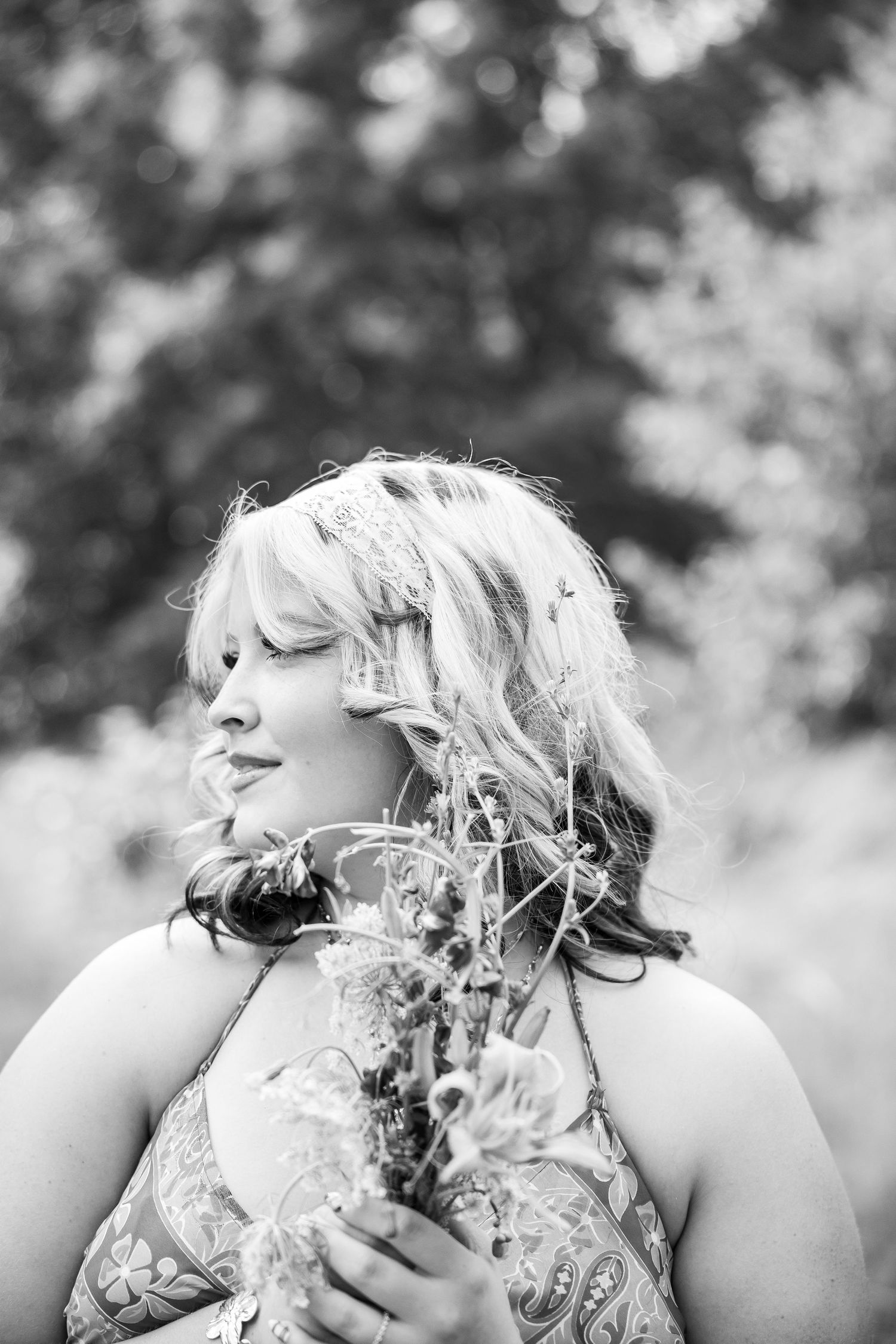Black and white portrait of a blonde woman holding wildflowers outdoors, looking away dreamily.