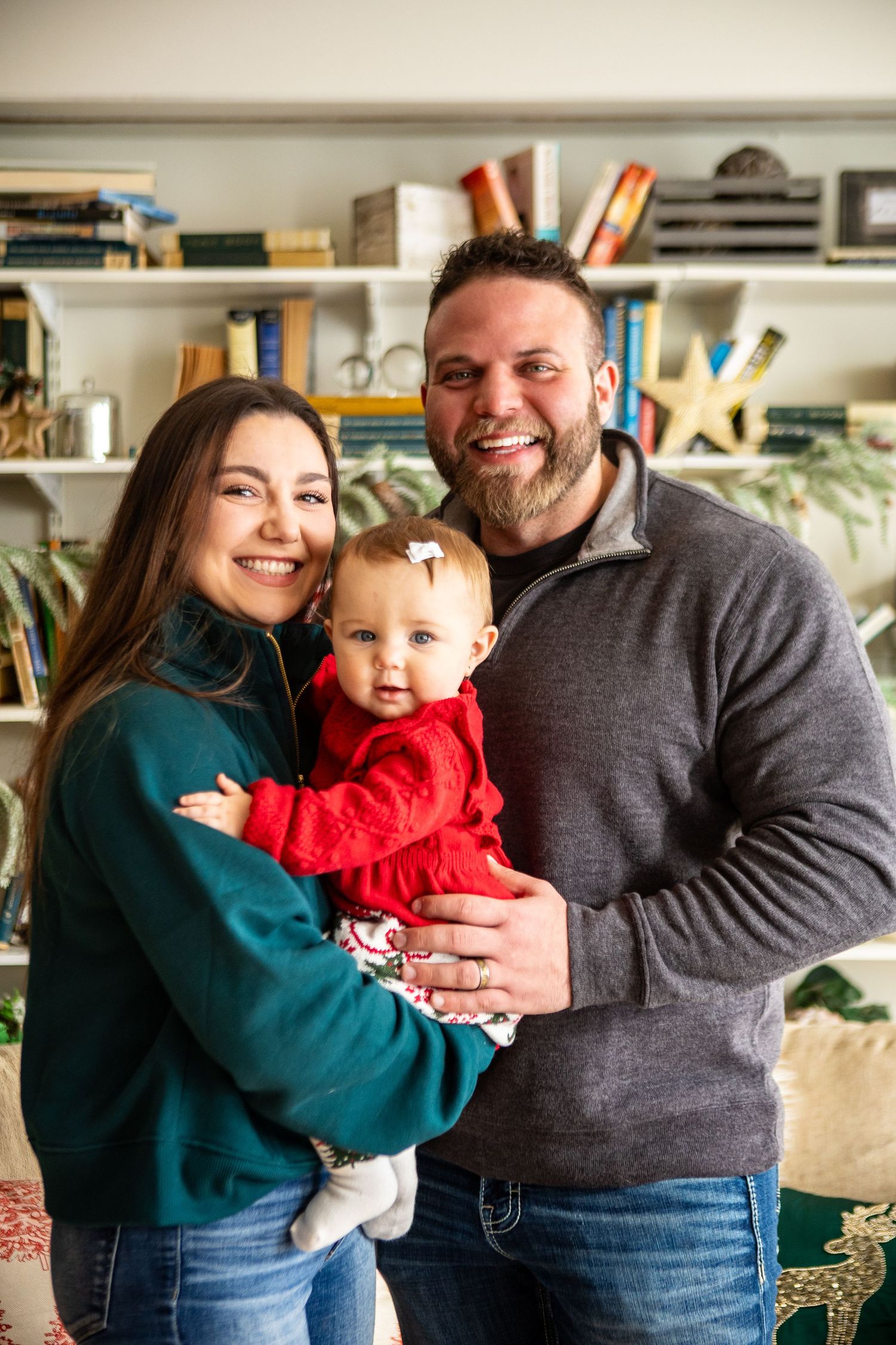 A family poses together in front of bookshelves, with a baby in a red sweater being held between two adults.