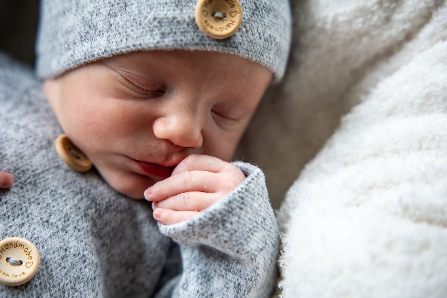 Sleeping newborn baby in gray knit outfit with wooden buttons, peacefully resting with tiny hand near face.